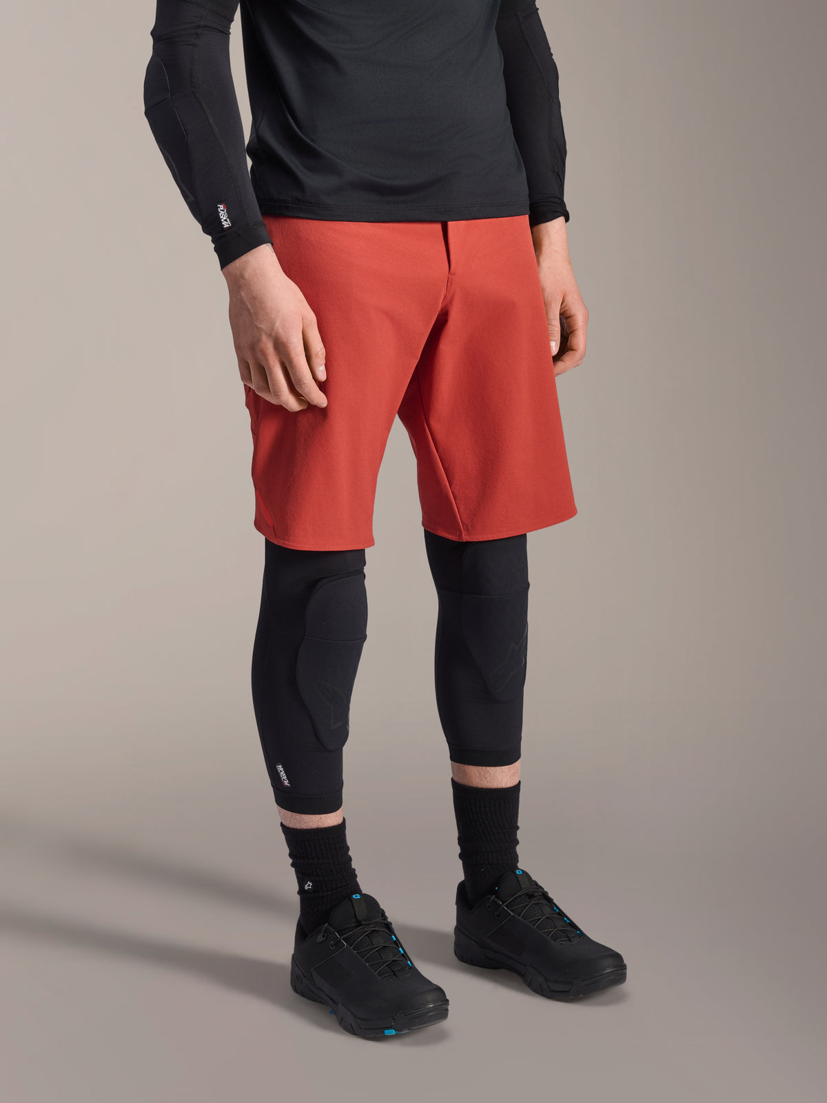 Alpinestars A-Motion Plasma Kniebandage, MTB-Knieschützer, Schwarz, getragen mit roten Shorts und schwarzen Mountainbike-Schuhen, flaches Slip-On-Design mit konturierten Schutzpolstern und atmungsaktivem Mesh.