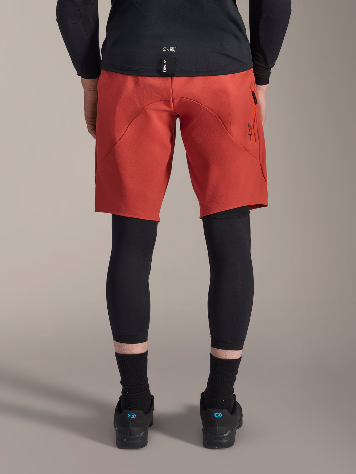 Alpinestars A-Motion Plasma Knie-Sleeve, schwarze MTB-Knieschützer, Rückansicht getragen unter roten Mountainbike-Shorts mit schwarzem Trikot und technischen Radschuhen, Schutzausrüstung für das Mountainbiken