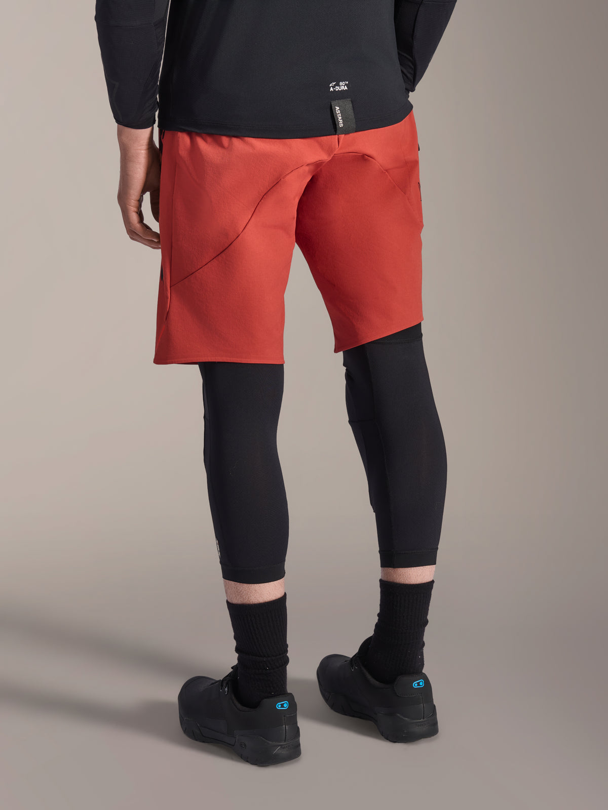 Alpinestars A-Motion Plasma Knieschützer, schwarzer MTB-Knieprotektor, getragen mit roten Mountainbike-Shorts und schwarzen Fahrradschuhen, Rückansicht des flachen Slip-on-Designs für den Offroad-Radsport