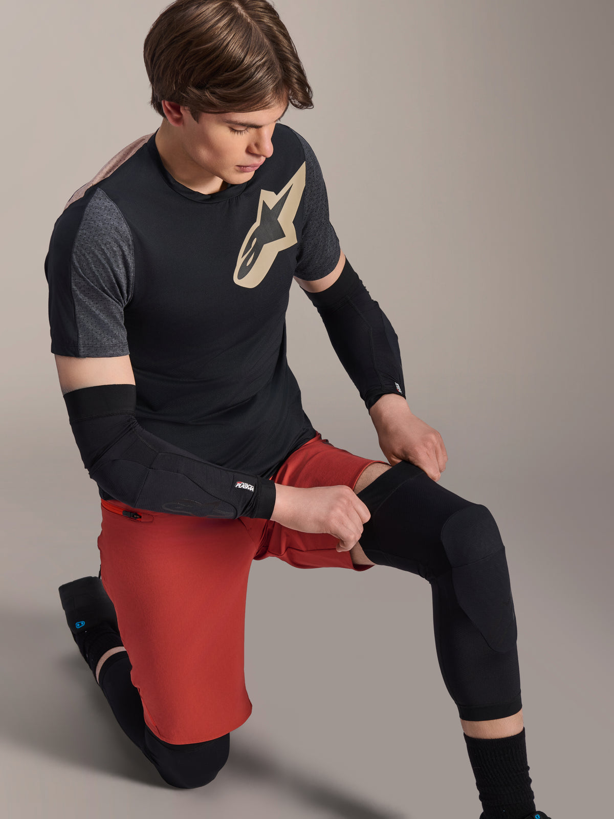 Alpinestars A-Motion Plasma Knie-Sleeve, MTB-Knieschützer, Schwarz, getragen von einem Fahrer in kniender Position, kombiniert mit einem schwarz-grauen T-Shirt mit goldenem Logo, roten Shorts und schwarzen Ellbogenschützern, professionelle Mountainbike-Schutzausrüstung
