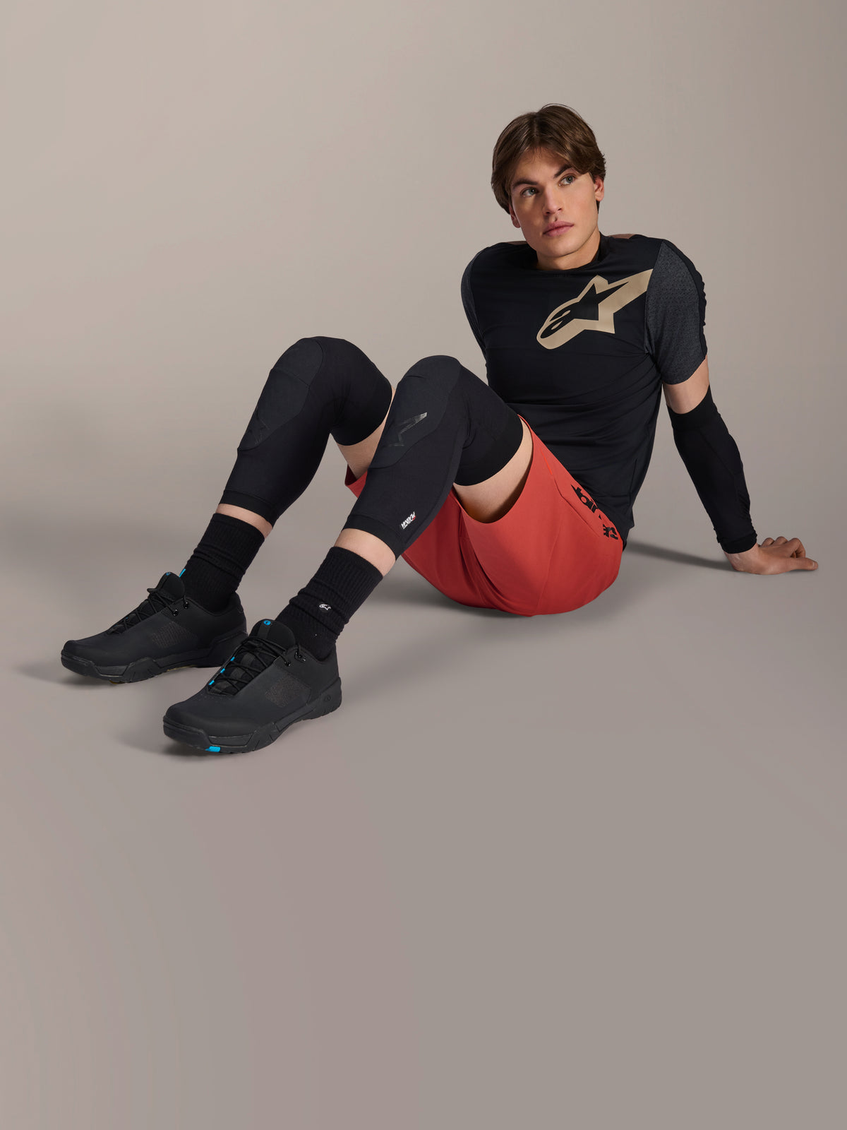 Alpinestars A-Motion Plasma Knee Sleeve, schwarze Mountainbike-Knieschützer, getragen von einem männlichen Model in kompletter Fahrradausrüstung mit roten Shorts und schwarzem Performance-T-Shirt, präsentiert das flache Schutzdesign.