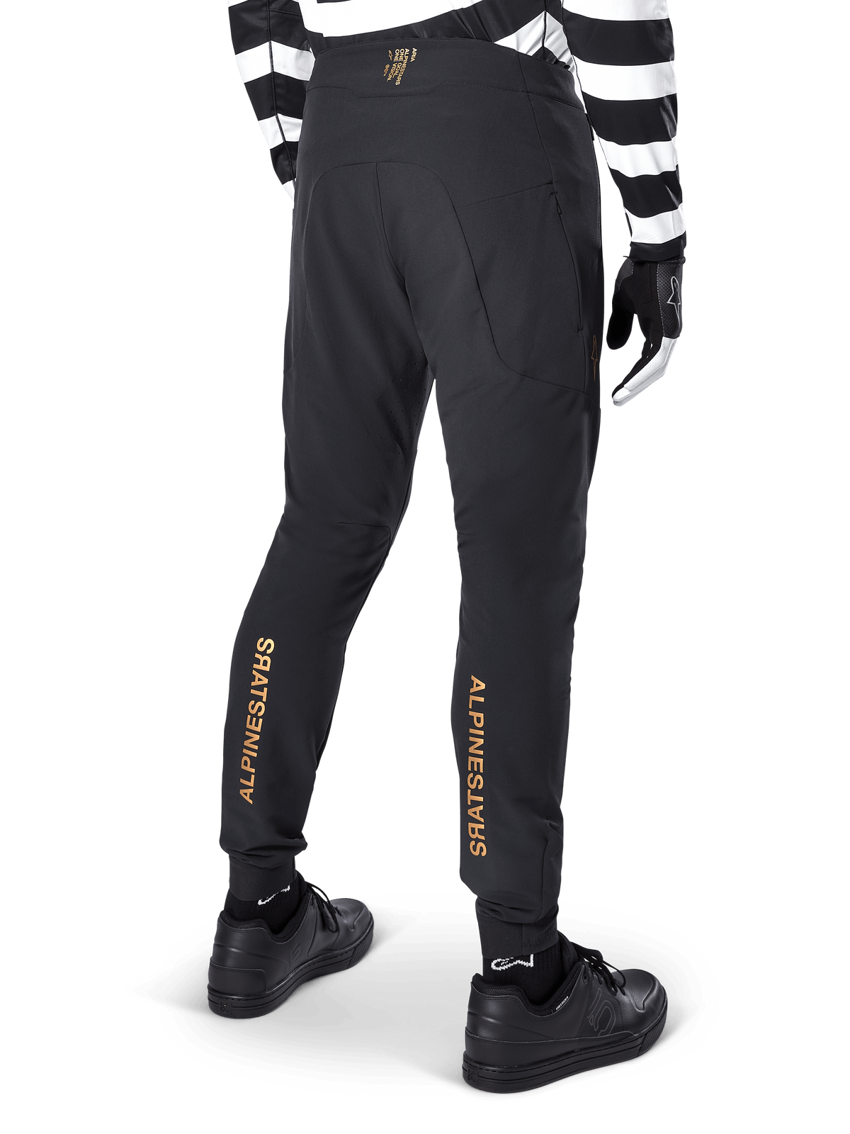 Alpinestars A-Aria Elite Pants, Black MTB Pants, rear view, featuring vertical gold Alpinestars branding on the back of the legs, worn with a black and white striped jersey and black shoes, performance cycling apparel for mountain biking