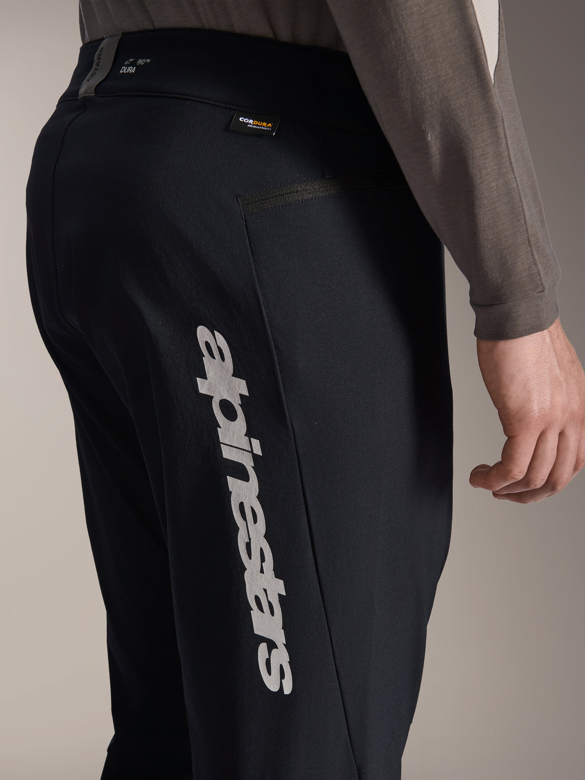 Alpinestars A-Dura Elite Hose, schwarze Mountainbike-Hose, Seitenansicht mit großem vertikalem Alpinestars-Logo am Bein, Cordura-Stoffetikett und seitlicher Reißverschlusstasche, technische Radsportbekleidung für Offroad-Fahrten