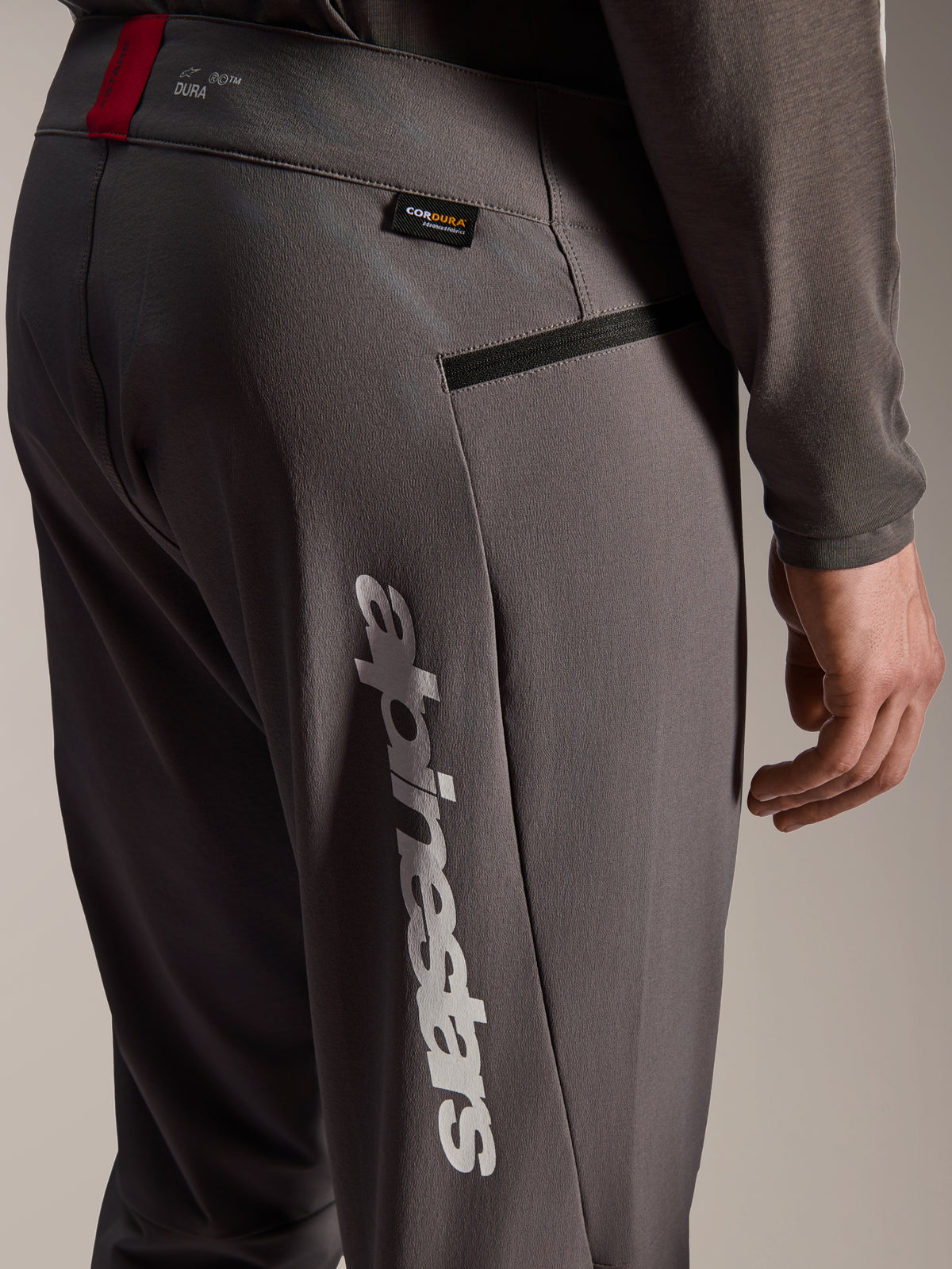 Alpinestars A-Dura Elite Hose, dunkelgraue MTB-Hose, Nahaufnahme von Hüfte und seitlichem Oberschenkel, mit großem weißem vertikalem Alpinestars-Logo, Cordura-Stoffetikett, Reißverschlusstasche und rotem Akzent am Bund
