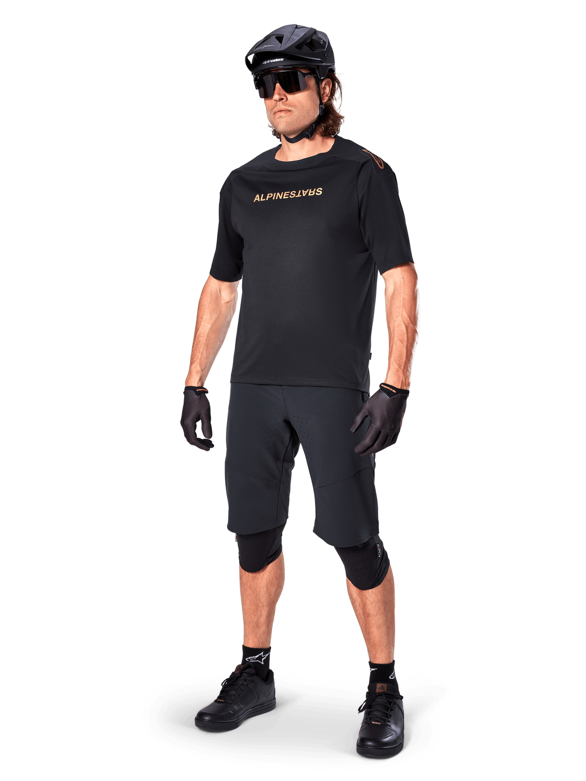Cyclist wearing Alpinestars A-Aria Elite Shorts, Black Mountain Bike Shorts, paired with a matching black jersey with gold branding, black helmet, sunglasses, and knee pads, complete performance outfit for off-road cycling