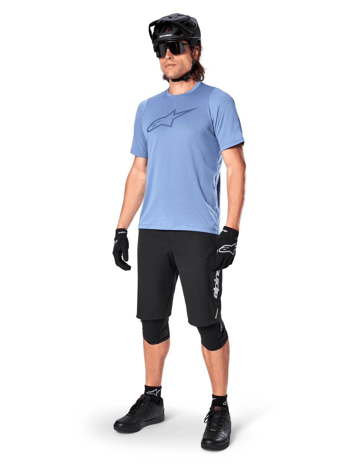 Männlicher Fahrer in Alpinestars A-Dura Elite Shorts in Schwarz, hellblaues kurzärmeliges Trikot mit Logo auf der Brust, schwarzer Helm, Sonnenbrille und schützende Knieschoner, komplette Mountainbike-Ausrüstung für Offroad-Trails.