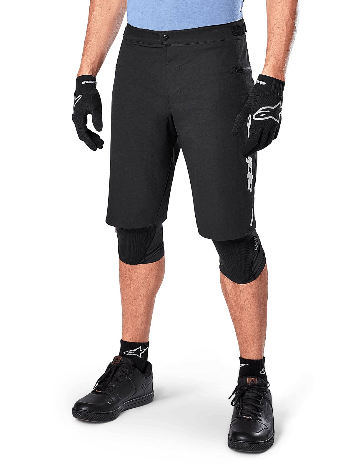 Alpinestars A-Dura Elite Shorts, schwarze MTB-Mountainbike-Shorts, getragen mit hellblauem Shirt und schwarzem Knieschutz, mit sicherem Druckknopfverschluss und Reißverschlusstasche, kombiniert mit schwarzen Radschuhen und Handschuhen für ein komplettes Offroad-Outfit.
