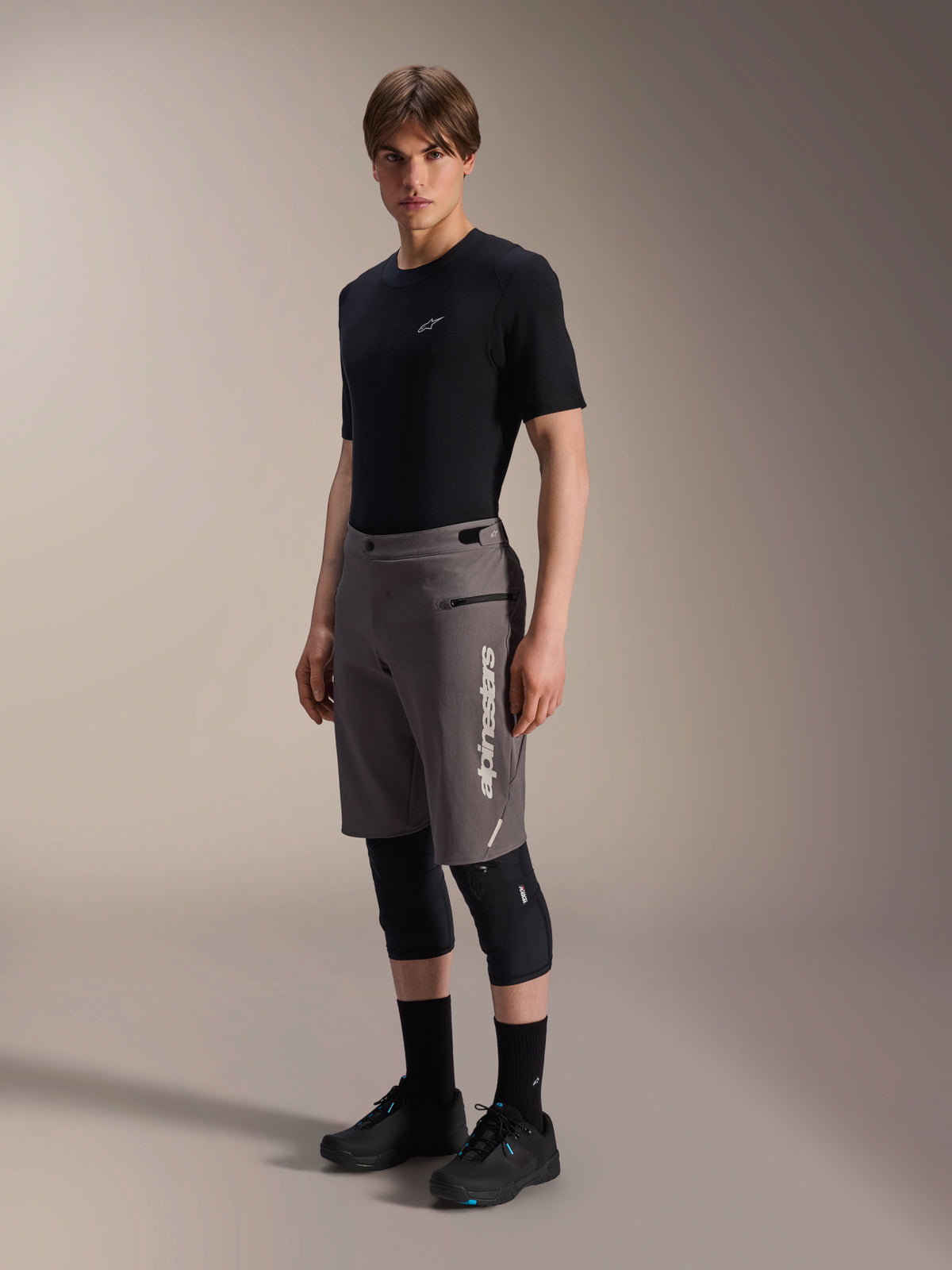 Alpinestars A-Dura Elite Shorts, dunkelgraue Mountainbike-Shorts, von einem Model mit schwarzem T-Shirt und Knieschützern getragen, mit vertikalem weißem Alpinestars-Logo auf dem linken Bein, technisches Cordura-Gewebe für Offroad-Radsport