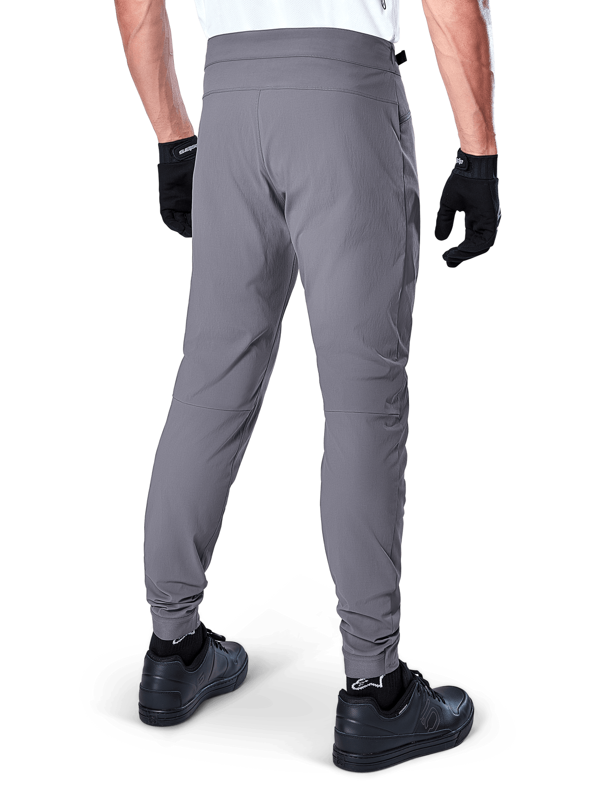 Alpinestars A-Dura Hose, Mountainbike-Hose, Dunkelgrau, Rückansicht eines Modells beim Tragen der Ausrüstung, mit strapazierfähigem 2-Wege-Stretch-Material und elastischen Bündchen, kombiniert mit schwarzen Handschuhen und Sneakern für einen radsporttauglichen Look