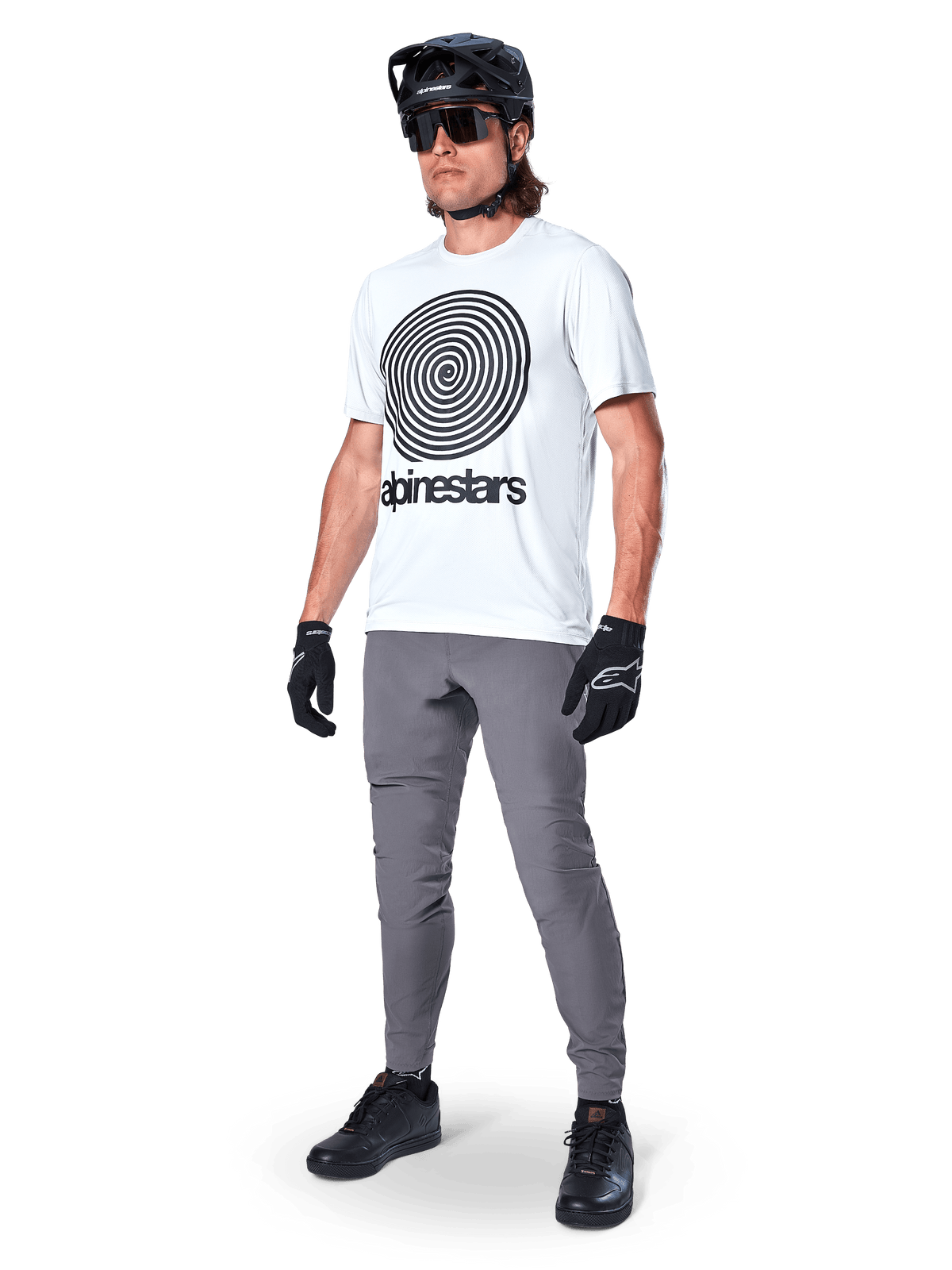 Alpinestars A-Dura Hose, MTB-Hose, Dunkelgrau, getragen mit weißem T-Shirt mit Spiralgrafik, schwarzem MTB-Helm, Sonnenbrille und schwarzen Handschuhen, strapazierfähiges 2-Wege-Stretch-Textil für den Offroad-Radsport