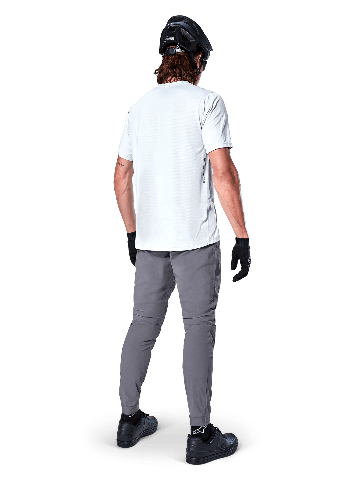 Alpinestars A-Dura Hose, dunkelgraue Mountainbike-Hose in der Rückansicht, getragen mit einem weißen Kurzarm-T-Shirt, schwarzem MTB-Helm, schwarzen Handschuhen und Schuhen, reguläre Passform mit elastischen Bündchen, strapazierfähige Offroad-Fahrradausrüstung