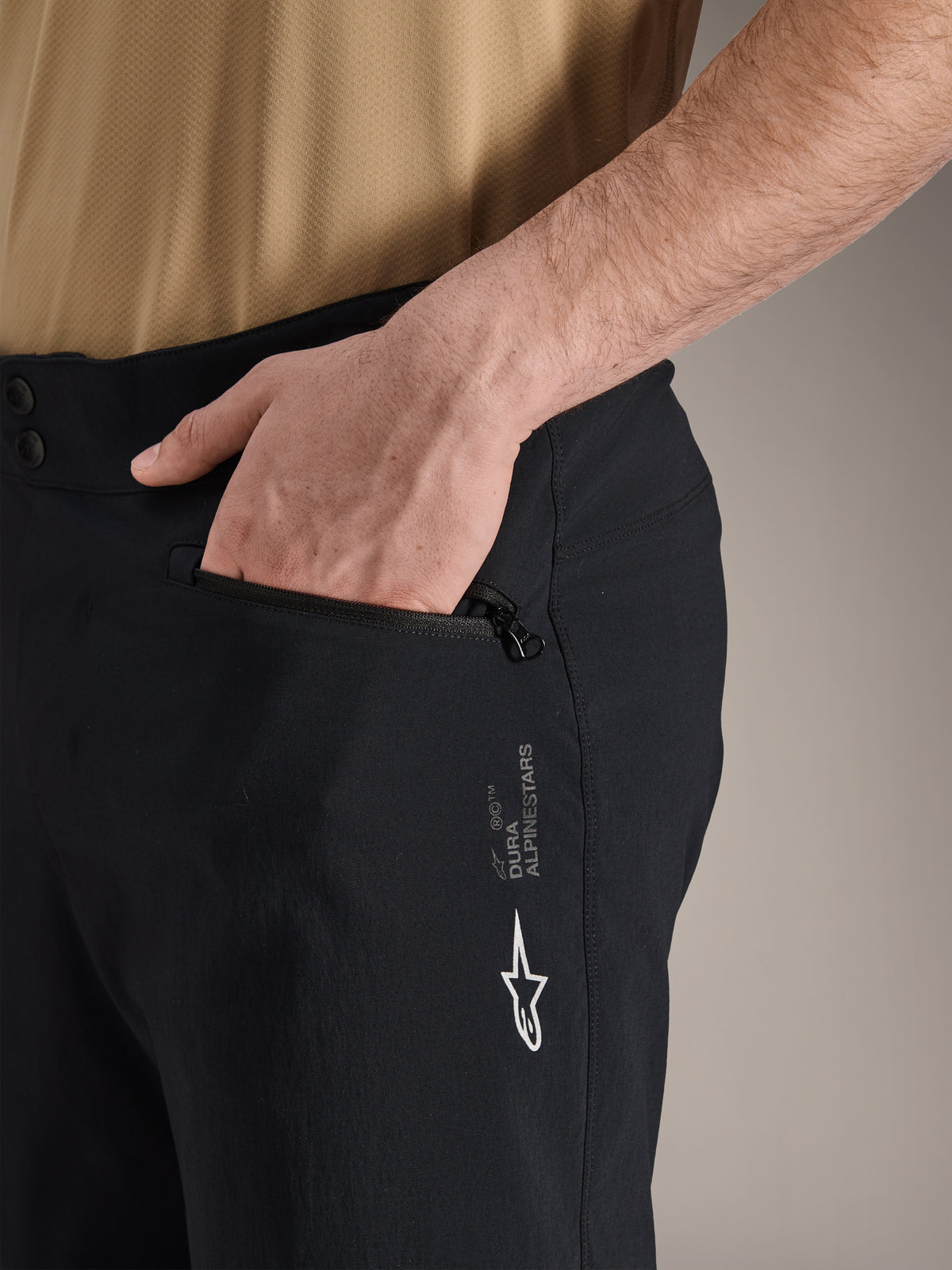 Alpinestars A-Dura Shorts, schwarze Mountainbike-Shorts, Nahaufnahme einer seitlichen Reißverschlusstasche mit Hand darin, mit weißem Alpinestars-Logo und A-Dura-Branding, strapazierfähiges Textil für den Offroad-Radsport