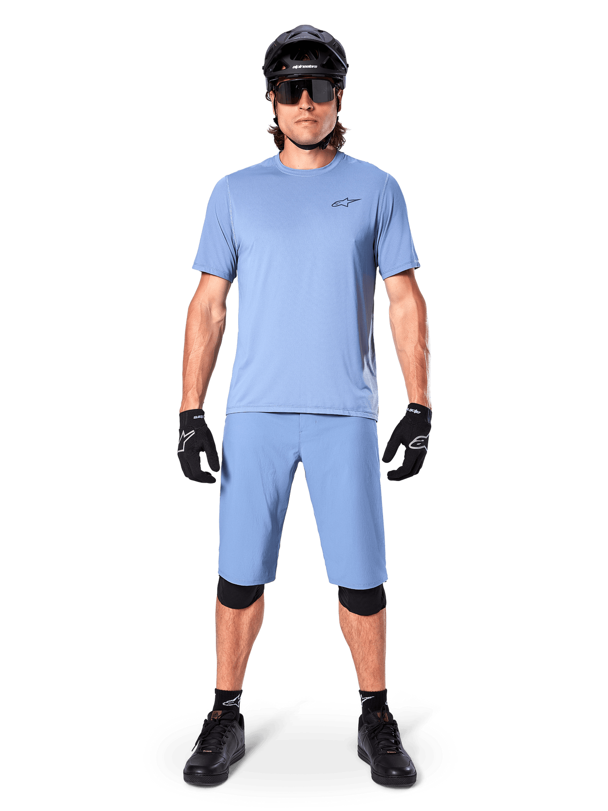 Model wearing Alpinestars A-Dura Shorts in Infinity Blue, matching light blue jersey, black mountain bike helmet, black sunglasses, black gloves and knee pads, complete MTB outfit for trail riding