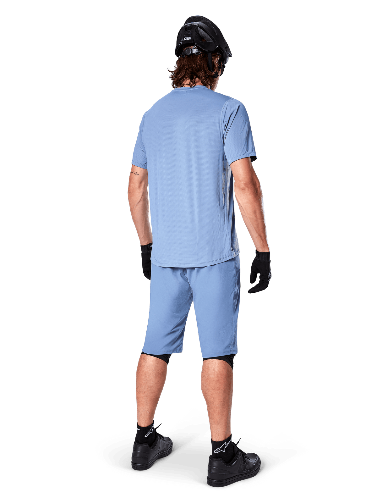Rear view of a mountain biker wearing Alpinestars A-Dura Shorts in Infinity Blue, matching short-sleeve jersey, black helmet, black gloves, and black shoes, coordinated performance gear for MTB trail riding