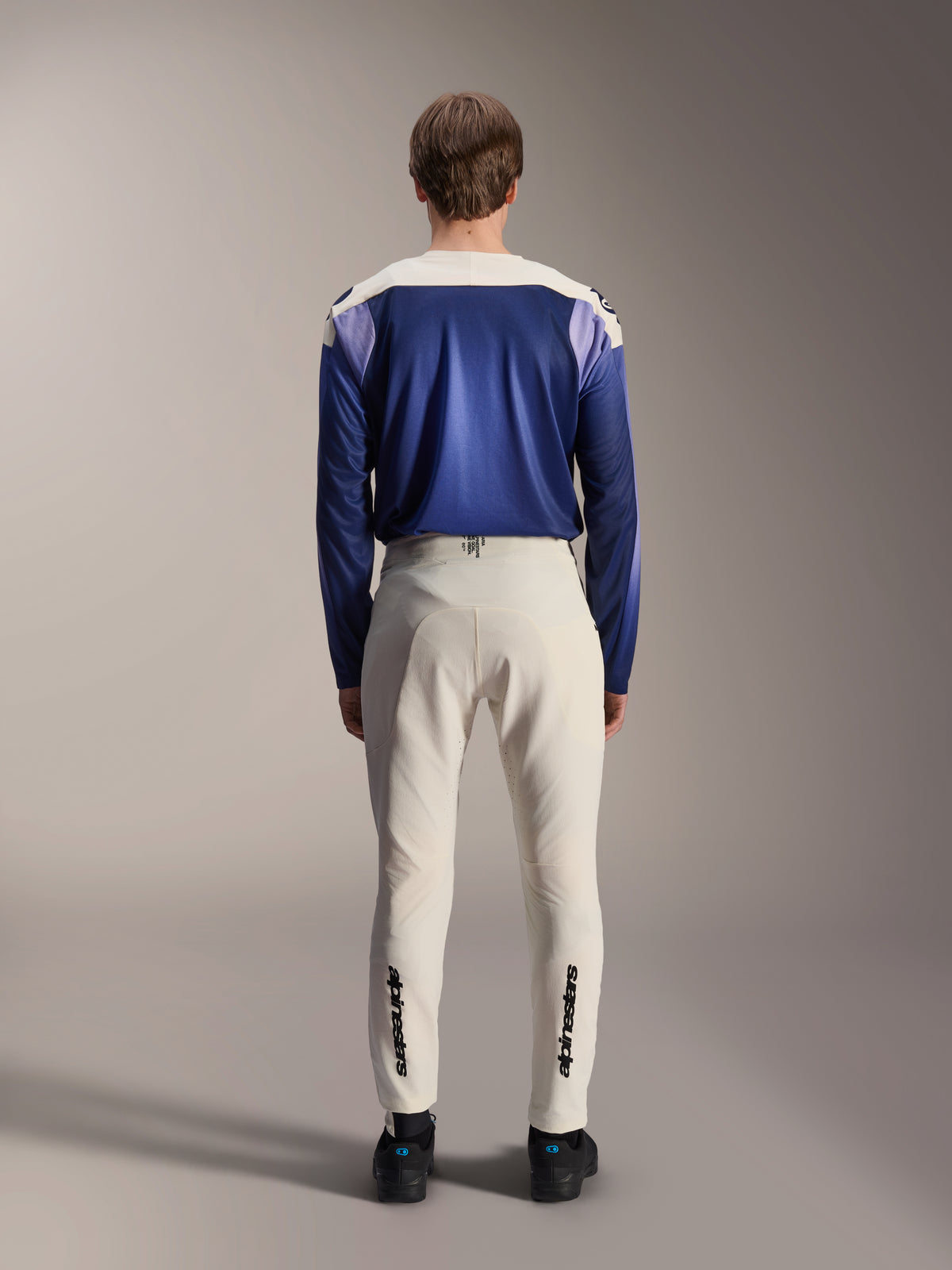 Alpinestars A-Aria Elite Pants in Ecru, shown from the back on a model paired with a blue A-Aria Polartec Maker Elite Long Sleeve Jersey, off-white technical MTB pants featuring black logos on the lower legs, performance cycling gear for mountain biking