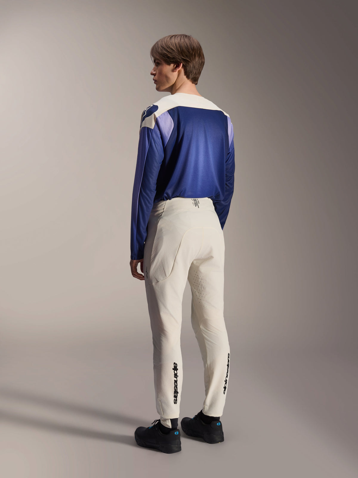 Alpinestars A-Aria Polartec Maker Elite Long Sleeve Jersey, Baja Blue, worn by a male model from behind with off-white MTB pants and black shoes, featuring a blue and white color-blocked design, performance mountain bike gear