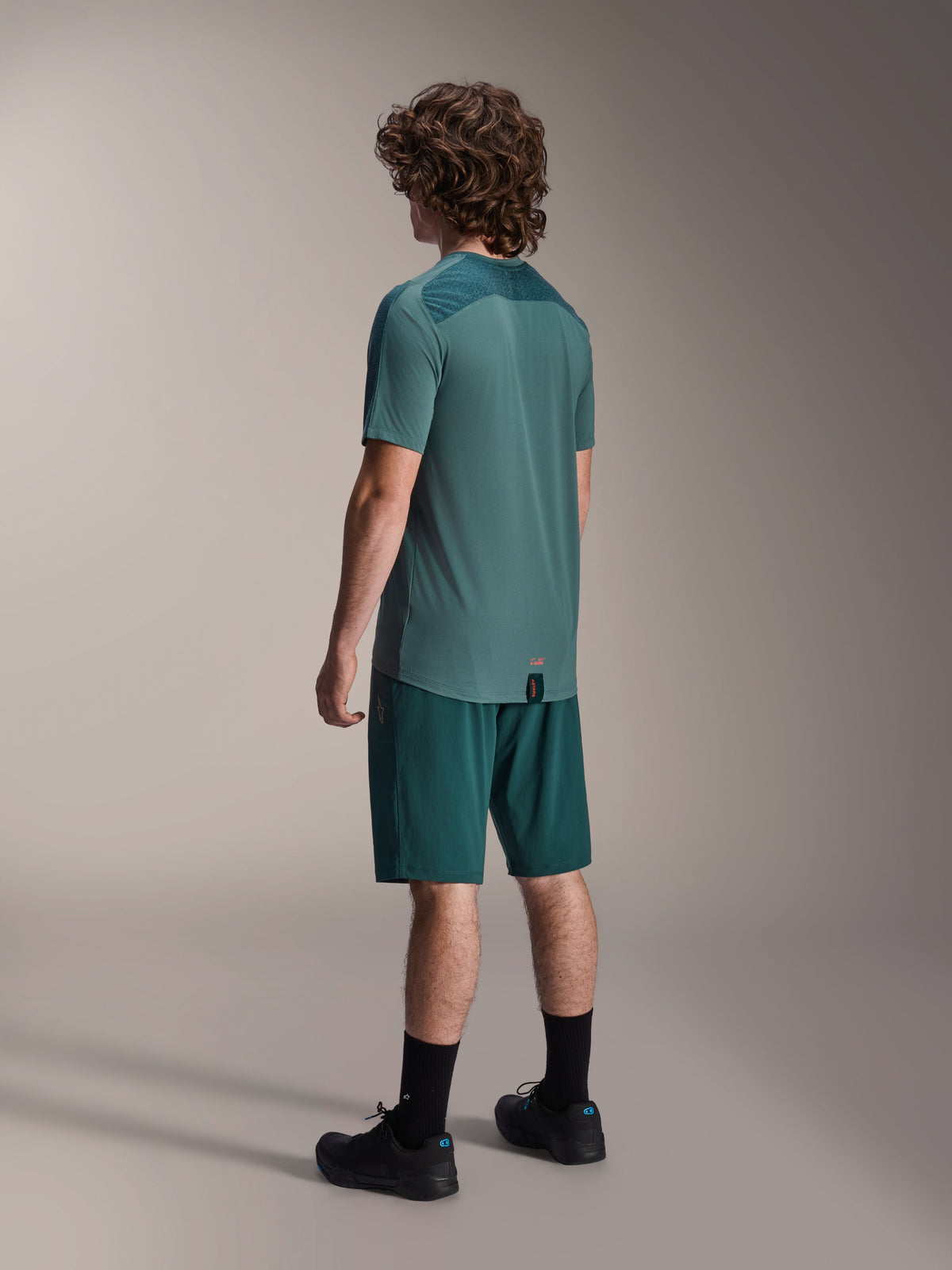 Male model wearing the Alpinestars A-Dura Lipan Short Sleeve Jersey in Silver Pine, seen from the back, paired with matching dark green mountain bike shorts, black socks and shoes, featuring a textured upper back panel and a dropped tail hem for extra coverage.
