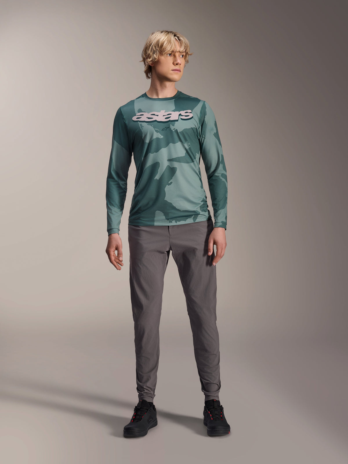 Alpinestars A-Dura Hollow Camo Langarm-Trikot, Silver Pine Camo mit grünem Camouflage-Muster, großes weißes „astars“-Logo auf der Brust, getragen mit grauer technischer Hose und schwarzen Radschuhen, Hochleistungs-MTB-Ausrüstung
