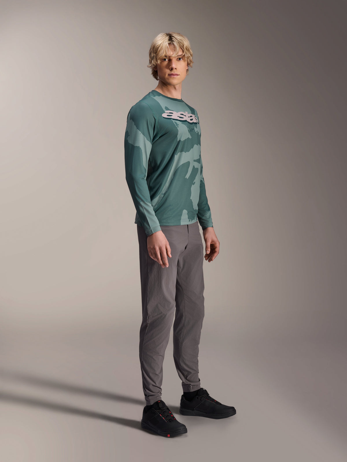 Alpinestars A-Dura Hollow Camo Langarm-Trikot, Silver Pine Camo, getragen von einem Model mit grauer Hose und schwarzen Schuhen, mit grünem Camouflage-Muster und markantem weißen ASTARS-Logo auf der Brust, Hochleistungs-MTB-Bekleidung