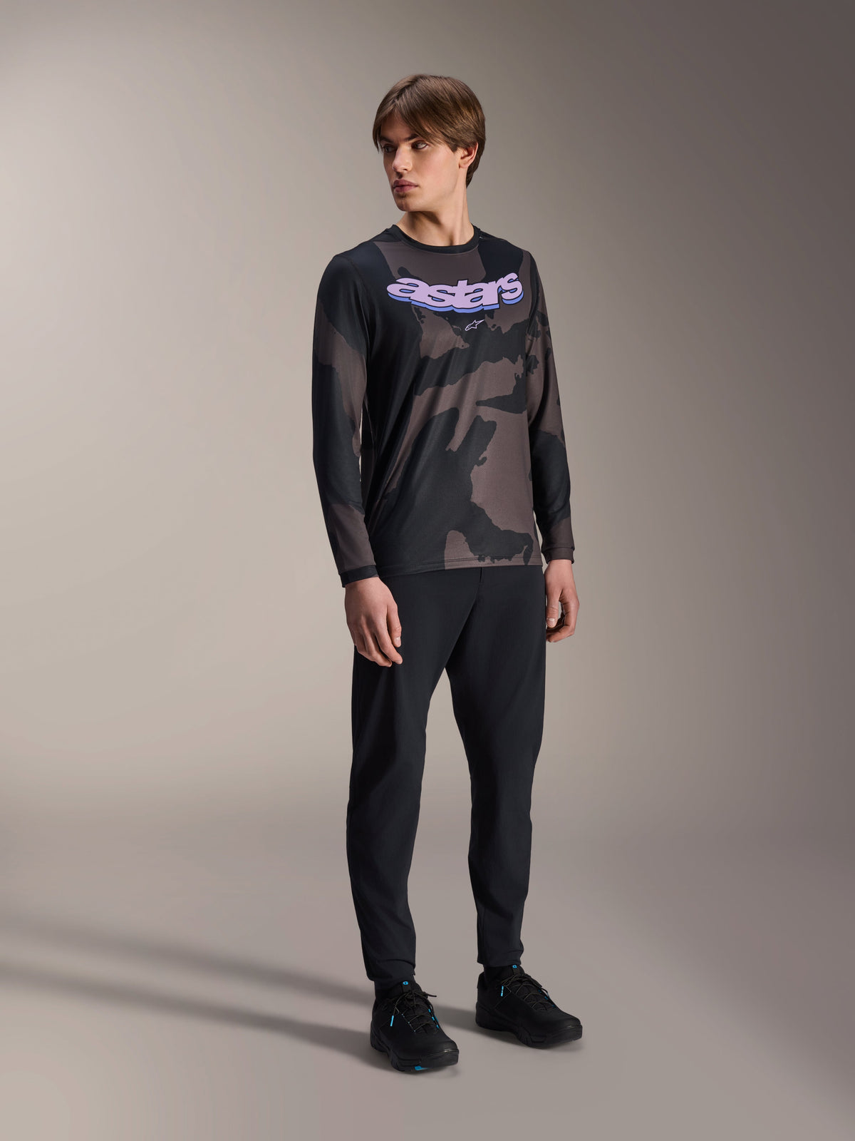 Männliches Modell im Alpinestars A-Dura Hollow Camo Langarm-Trikot, Black Camo Muster mit lilafarbener 'astars' Brustgrafik, kombiniert mit schwarzer technischer Hose und schwarzen Schuhen, lässige MTB-Ausrüstung