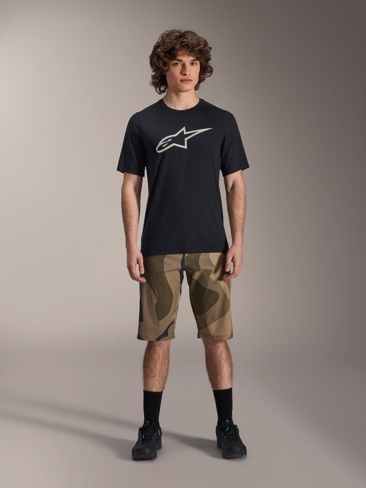 Alpinestars A-Dura Astar Kurzarm-Trikot, Schwarz mit großem weißem Astars-Logo auf der Brust, getragen von einem Model mit Camouflage-MTB-Shorts und schwarzen Radschuhen, lässige Mountainbike-Bekleidung.