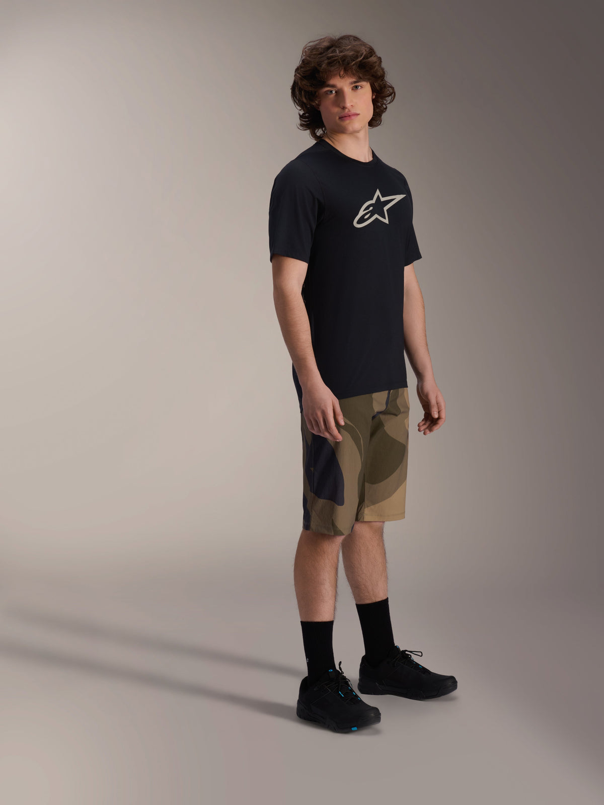 Alpinestars A-Dura Astar Kurzarm-Trikot, Schwarz mit weißem Brustlogo, getragen mit Camouflage-Shorts und schwarzen MTB-Schuhen, lässige Mountainbike-Ausrüstung