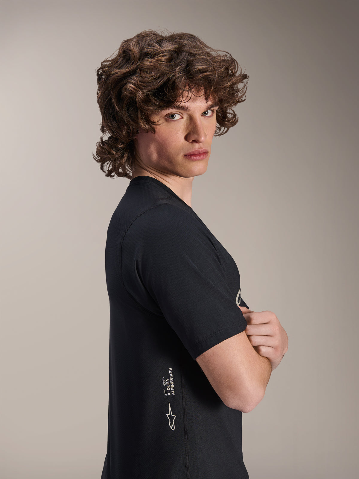 Alpinestars A-Dura Astar Jersey, Kurzarm-MTB-Trikot, Schwarz, männliches Model posiert im Profil mit verschränkten Armen, mit weißem Alpinestars-Branding an der unteren Seite, lässiges sportliches Design für Mountainbiking.