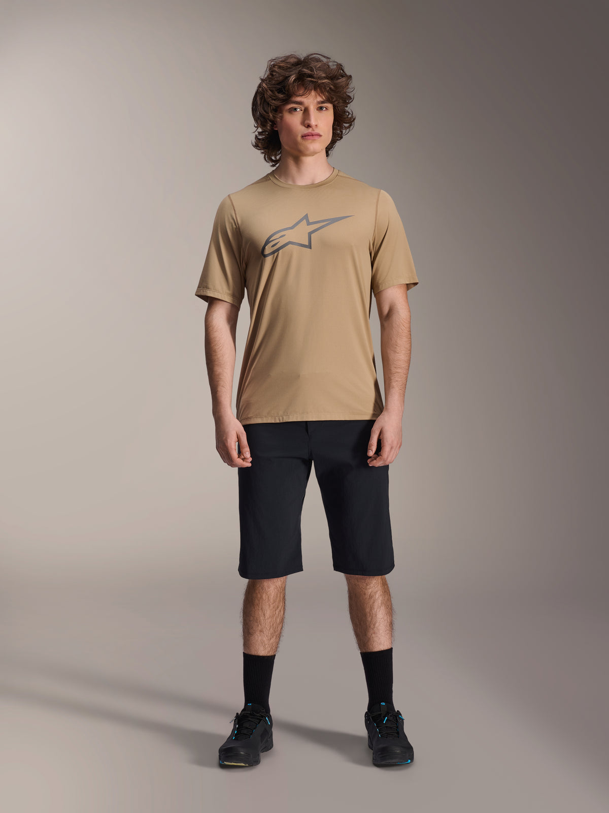 Model trägt Alpinestars A-Dura SS Jersey in Hellbraun und A-Dura Shorts in Schwarz, mit grauem Stern-Logo auf der Brust, kombiniert mit schwarzen Socken und Mountainbike-Schuhen, komplette MTB-Fahrradbekleidung in einer Studioaufnahme.