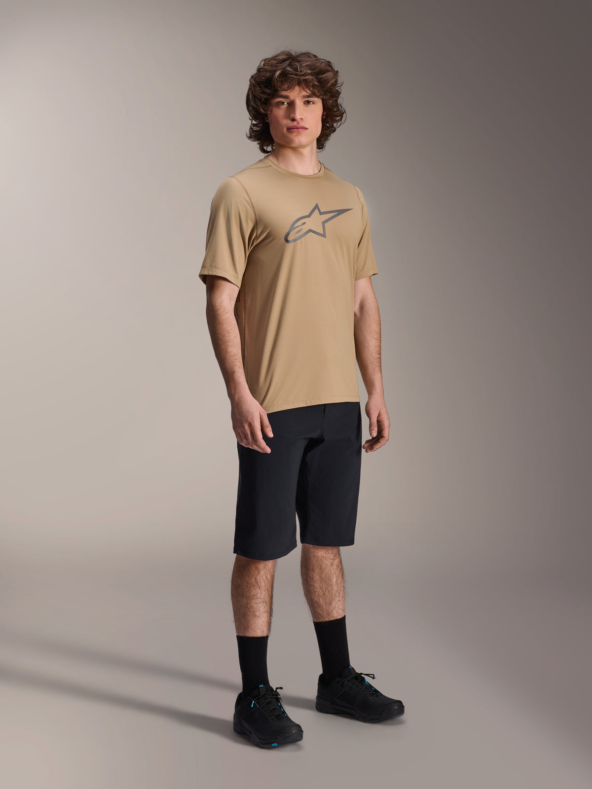 Alpinestars A-Dura Shorts, schwarze Mountainbike-Shorts, getragen von einem Model mit einem hellbraunen kurzärmeligen Trikot mit Alpinestars-Logo, schwarzen Socken und schwarzen Schuhen, strapazierfähige MTB-Ausrüstung für Geländefahrten