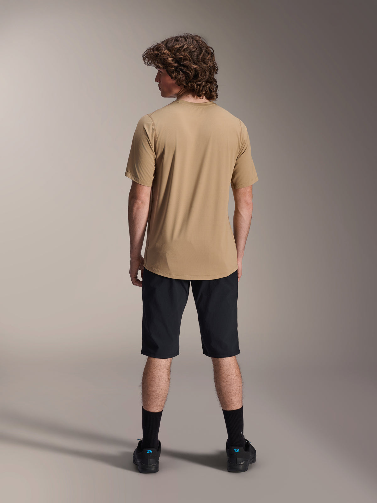 Männliches Model von hinten in schwarzen Alpinestars A-Dura Shorts und einem hellbraunen A-Dura Kurzarmtrikot, kombiniert mit schwarzen Socken und Mountainbikeschuhen, funktionales MTB-Trail-Outfit
