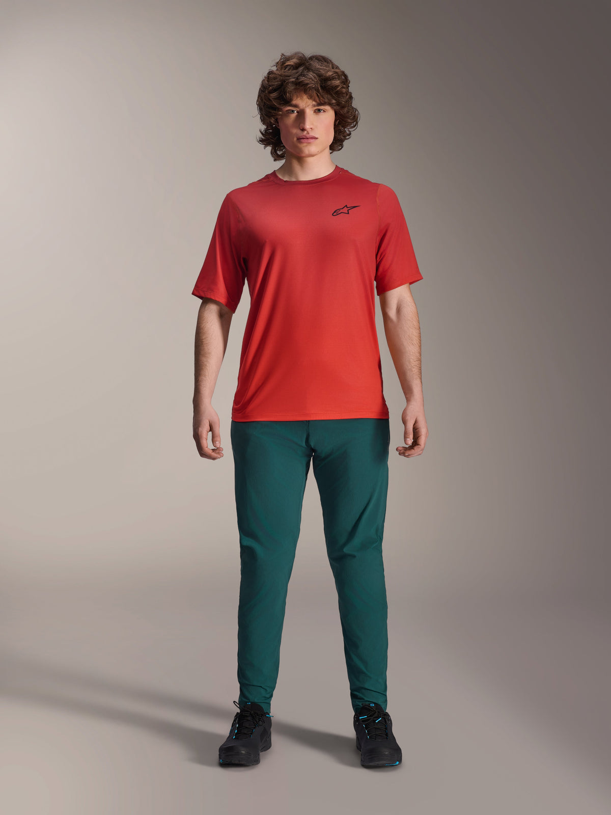 Alpinestars A-Dura Rise Kurzarmtrikot, Spicy Orange, getragen mit dunkelgrüner Hose und schwarzen Schuhen, mit schwarzem Astars-Logo auf der Brust, lässige Performance-Mountainbike-Bekleidung für Offroad-Fahrten