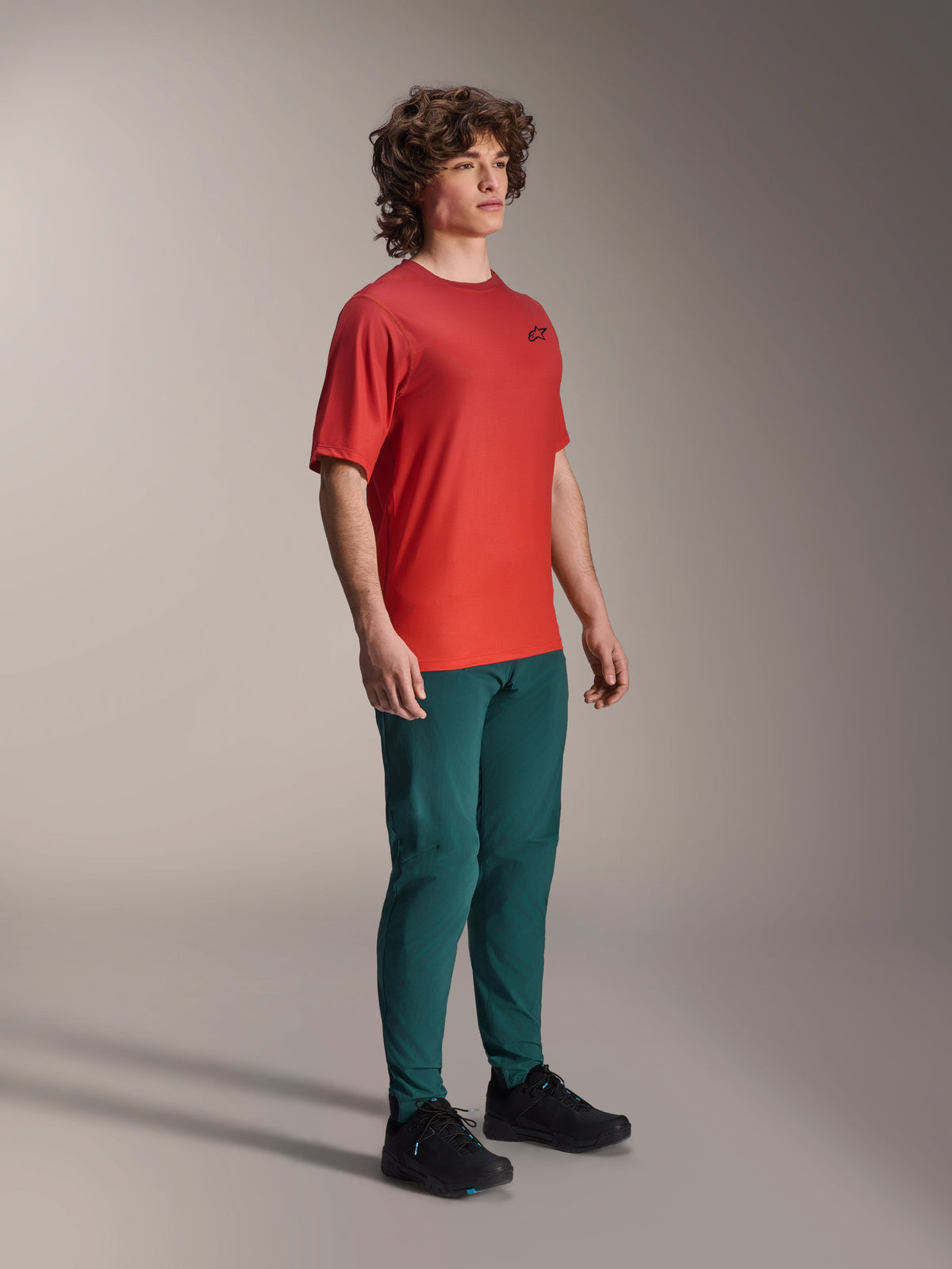 Alpinestars A-Dura Rise Kurzarmtrikot, Spicy Orange, mit kleinem schwarzen Brustlogo, getragen mit dunkel-petrolfarbener Hose und schwarzen Schuhen, leichte feuchtigkeitsableitende Mountainbike-Bekleidung.