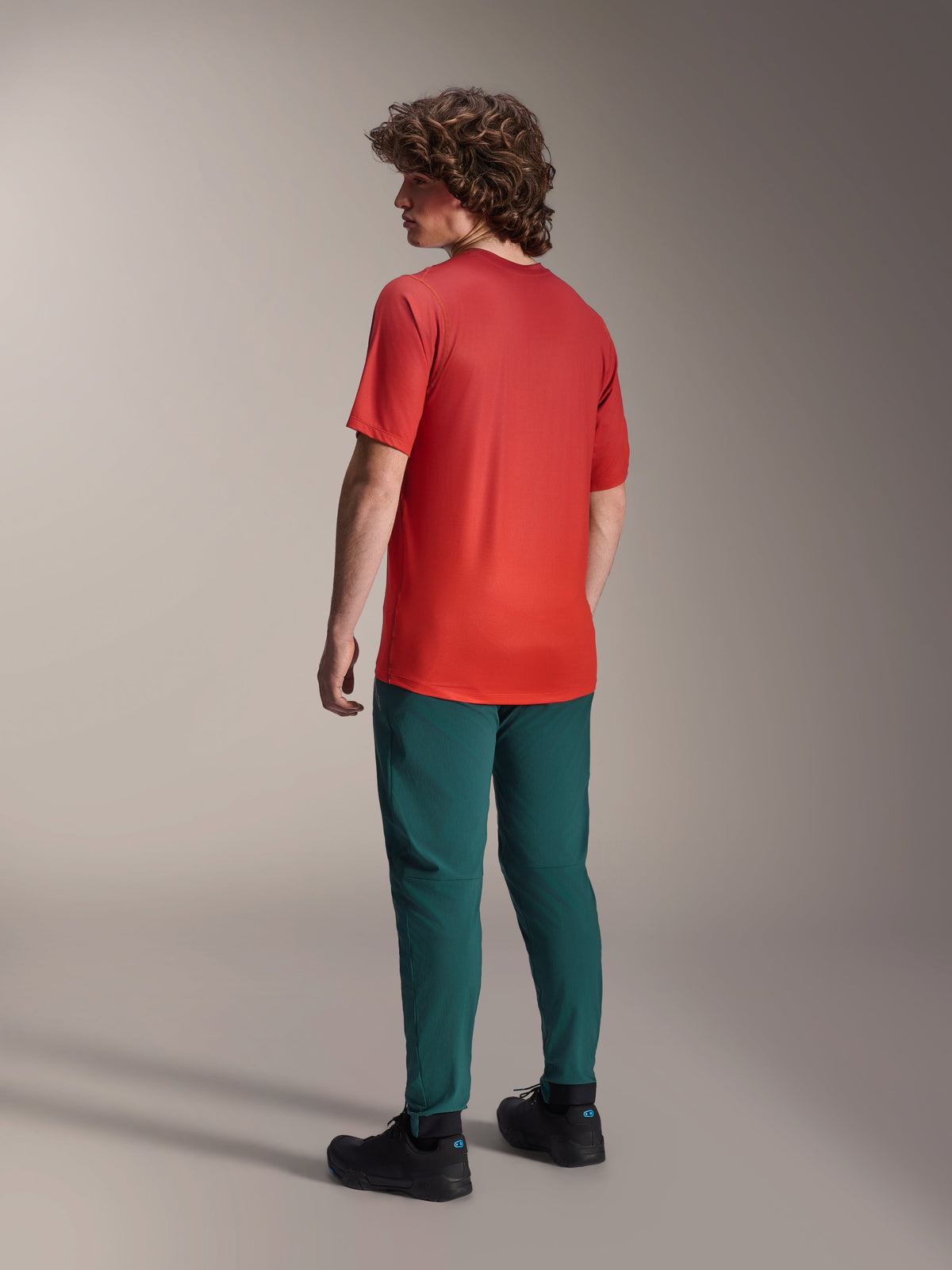 Alpinestars A-Dura Rise Kurzarmtrikot in Spicy Orange, Rückansicht eines Models mit dem Radtrikot, dunkelgrüner Mountainbike-Hose und schwarzen Schuhen, mit längerem abfallendem Saum und Performance-Passform für Geländefahrten.