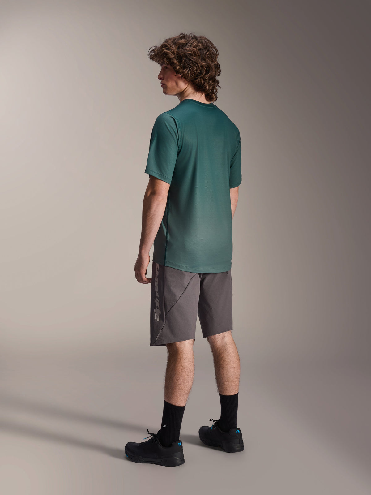 Alpinestars A-Dura Rise Trikot, Kurzarm-MTB-Trikot, Silver Pine Grün, getragen mit grauen Shorts und schwarzen Schuhen, Rückansicht mit verlängertem Rückensaum und regulärer Passform, leichtes, feuchtigkeitsableitendes Material für Trailfahrten