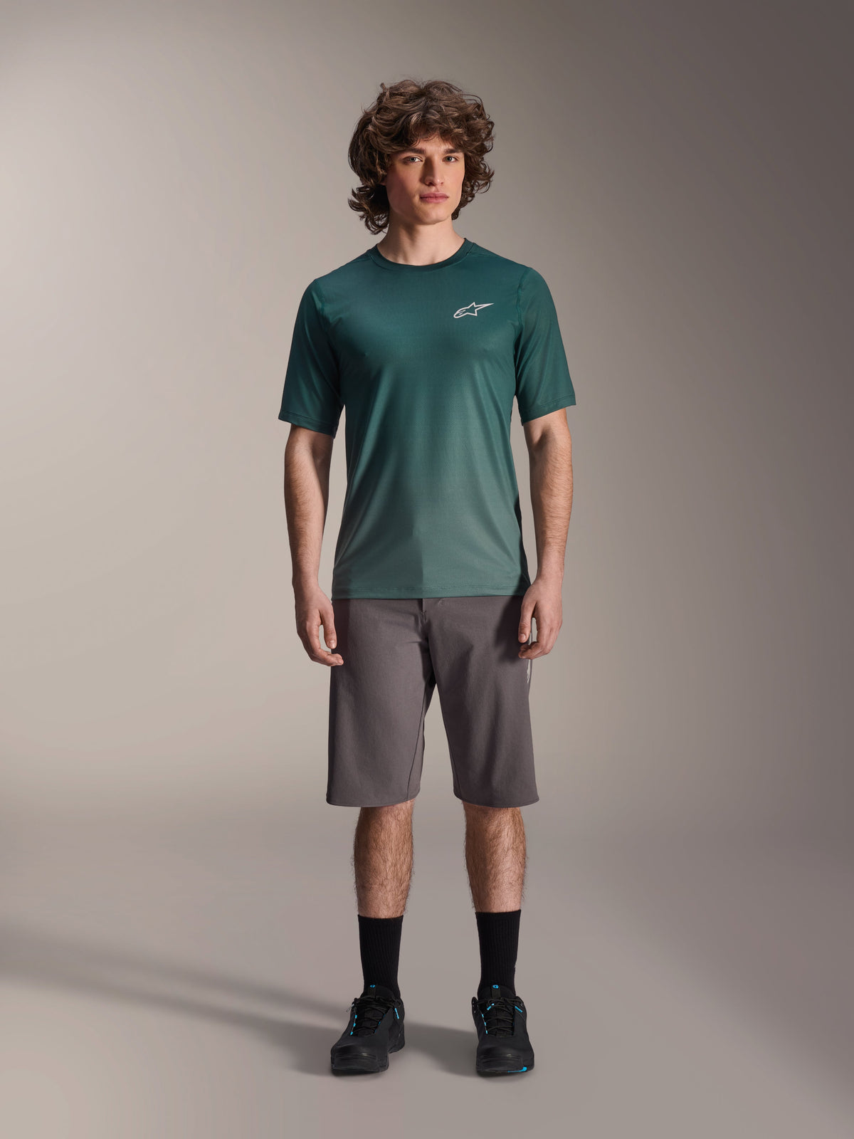 Männliches Model trägt Alpinestars A-Dura Rise Kurzarmtrikot in Silver Pine, grünes Radsporttrikot mit kleinem weißem Brustlogo, kombiniert mit grauen Mountainbike-Shorts und schwarzem Schuhwerk, lässige MTB-Trail-Ausrüstung
