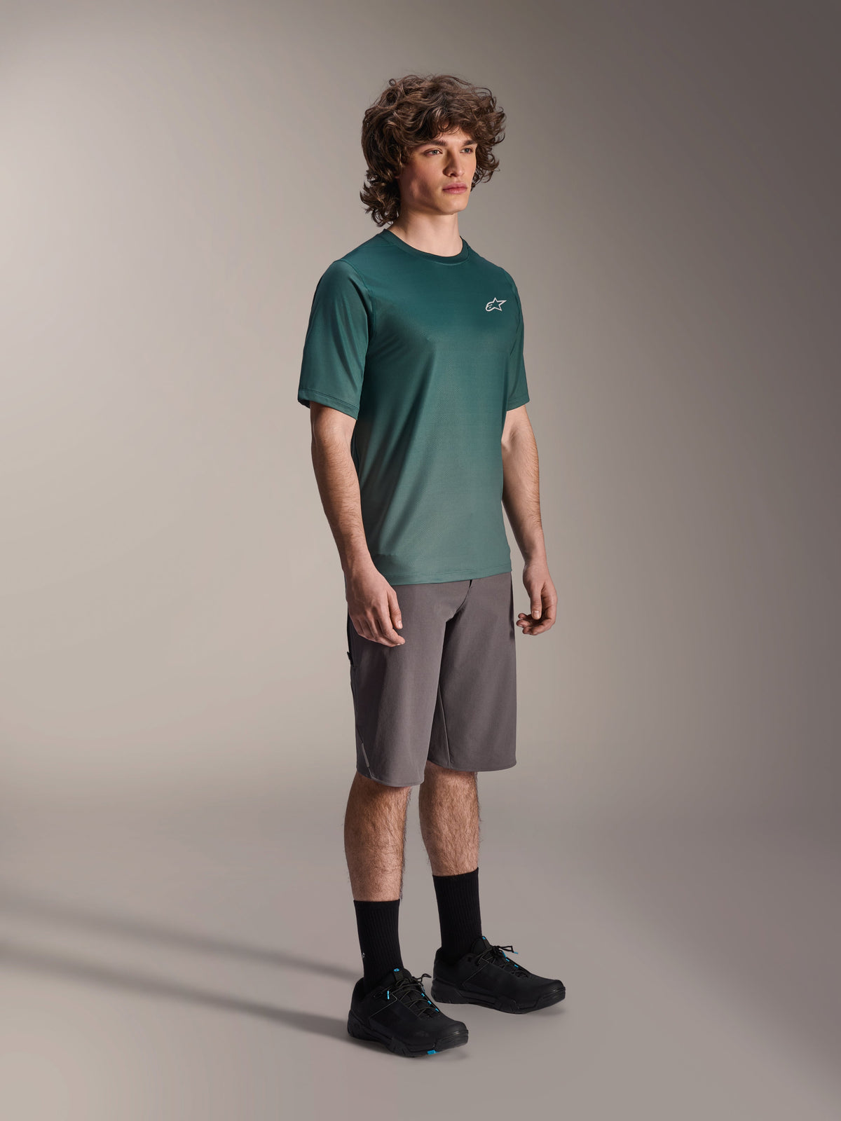 Alpinestars A-Dura Rise Kurzarmtrikot in Silver Pine, grünes Mountainbike-Trikot getragen von einem Model mit grauen Shorts und schwarzen Sneakern, lässige MTB-Trail-Ausrüstung mit kleinem weißen Logo auf der Brust