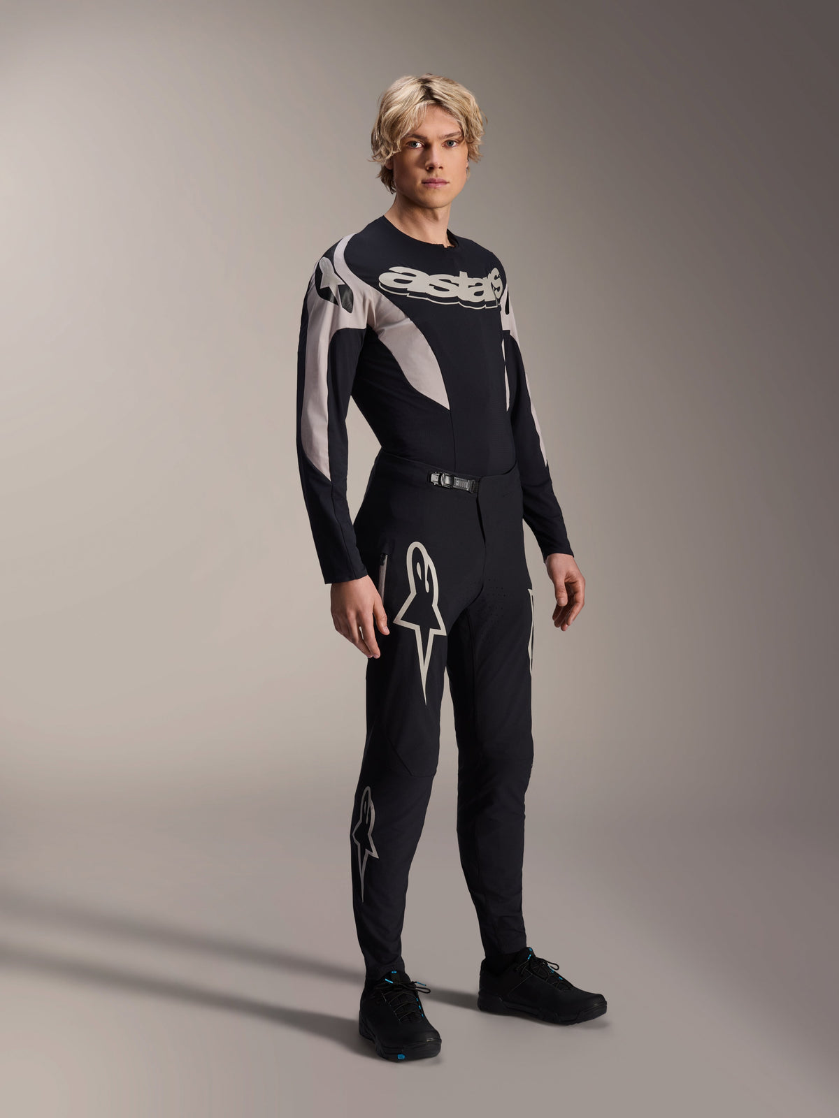 Männliches Model in Alpinestars A-Supra Melt Hose und passendem Langarm-Trikot, Schwarz mit hellgrauen geschwungenen Mustern, auffällige weiße Astars-Logos an den Beinen und Wortmarke auf der Brust, professionelle Mountainbike-Rennausrüstung.