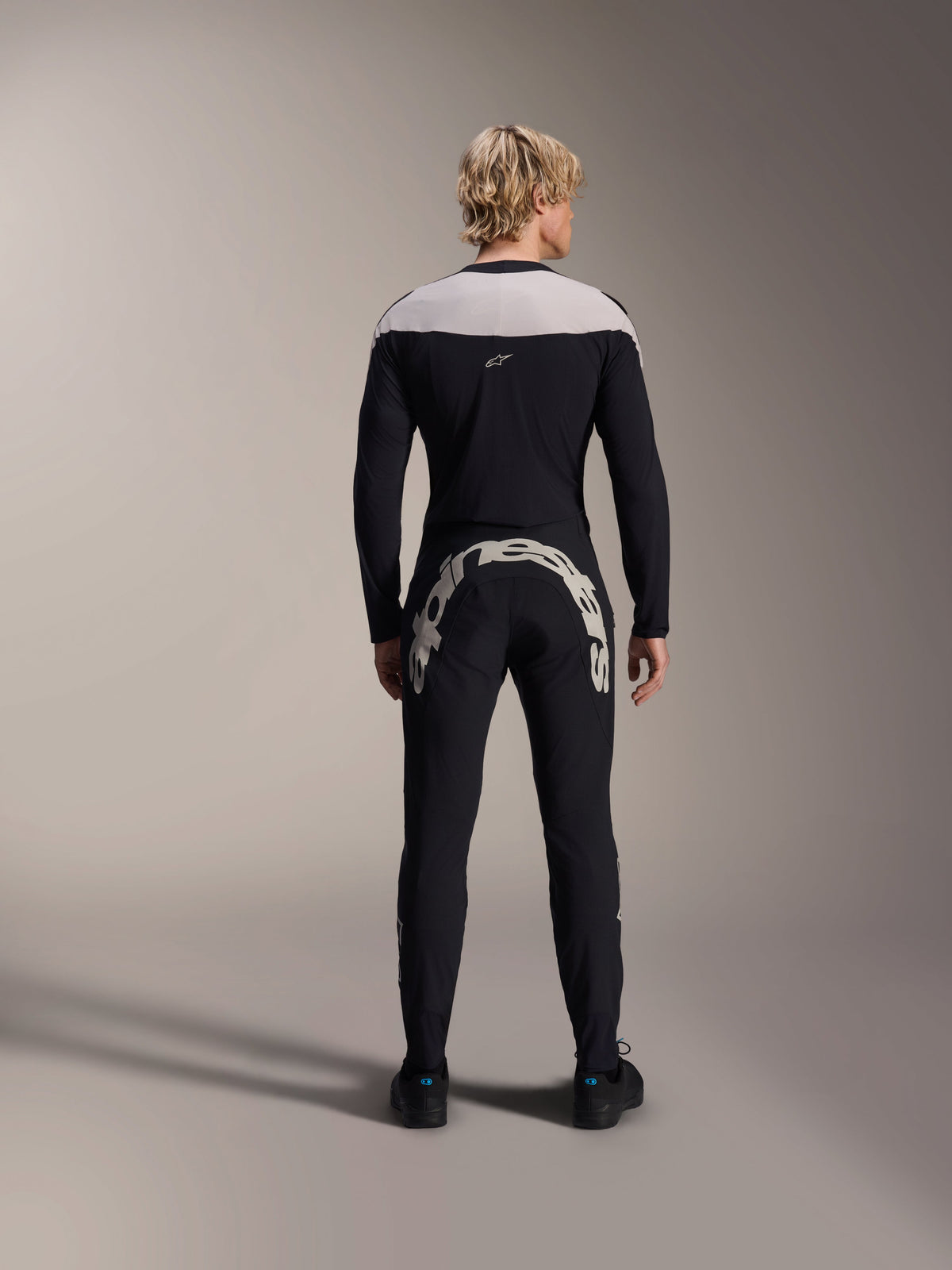 Alpinestars A-Supra Melt Hose, Schwarz, Rückansicht mit großem weißem Alpinestars-Schriftzug über dem hinteren Joch, kombiniert mit passendem Langarm-Trikot und schwarzen Radschuhen, Hochleistungs-Mountainbike-Ausrüstung für den Offroad-Einsatz.