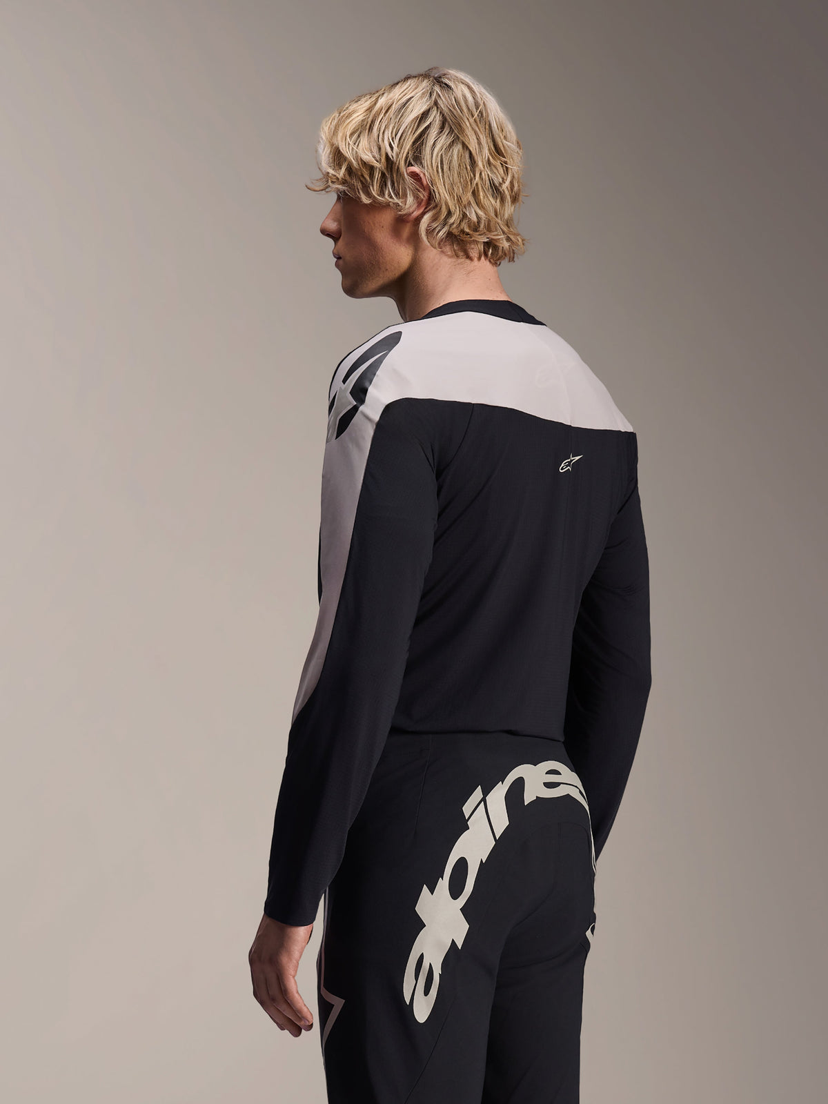 Männliches Model trägt das Alpinestars A-Supra Melt Langarmtrikot und die A-Supra Melt Hose, schwarz-hellgraue MTB-Rennausrüstung, mit großem, weißem Alpinestars-Schriftzug auf der Rückseite der Hose