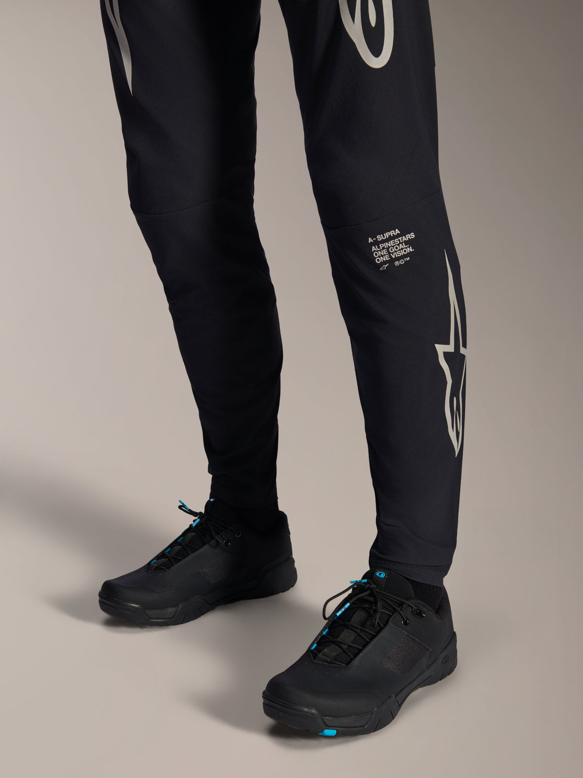 Alpinestars A-Supra Melt Hose, schwarze Mountainbike-Hose, weißer Alpinestars-Schriftzug und Logo auf dem Bein, schwarze Fahrradschuhe mit blauen Akzenten, Performance-Offroad-Ausrüstung