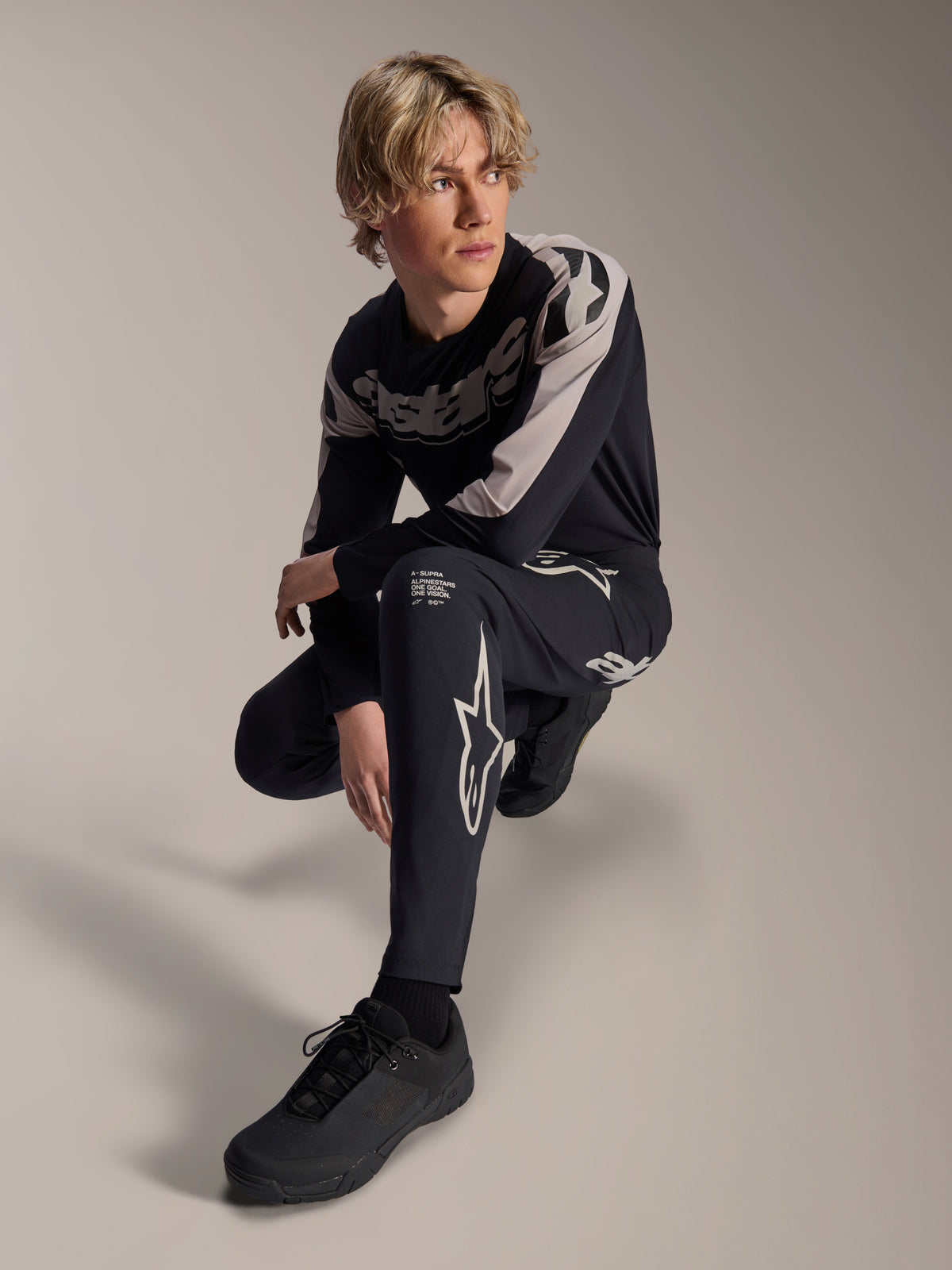 Alpinestars A-Supra Melt Hose und Langarmtrikot, Schwarz und Grau, mit auffälligem weißem Branding und Stern-Logos, getragen von einem männlichen Model in hockender Pose, abgestimmte MTB-Rennausrüstung