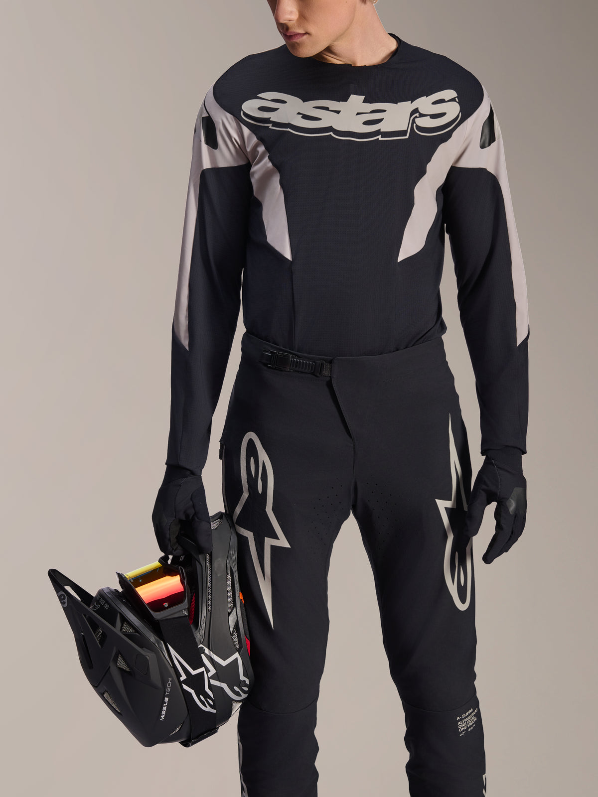 Alpinestars A-Supra Melt MTB-Racing-Kit, Langarmtrikot und Hose, Schwarz und Grau mit weißen Astars-Logos, Modell hält einen schwarzen Fullface-Helm und trägt passende Handschuhe, Hochleistungsausrüstung für Downhill- und Enduro-Mountainbiking