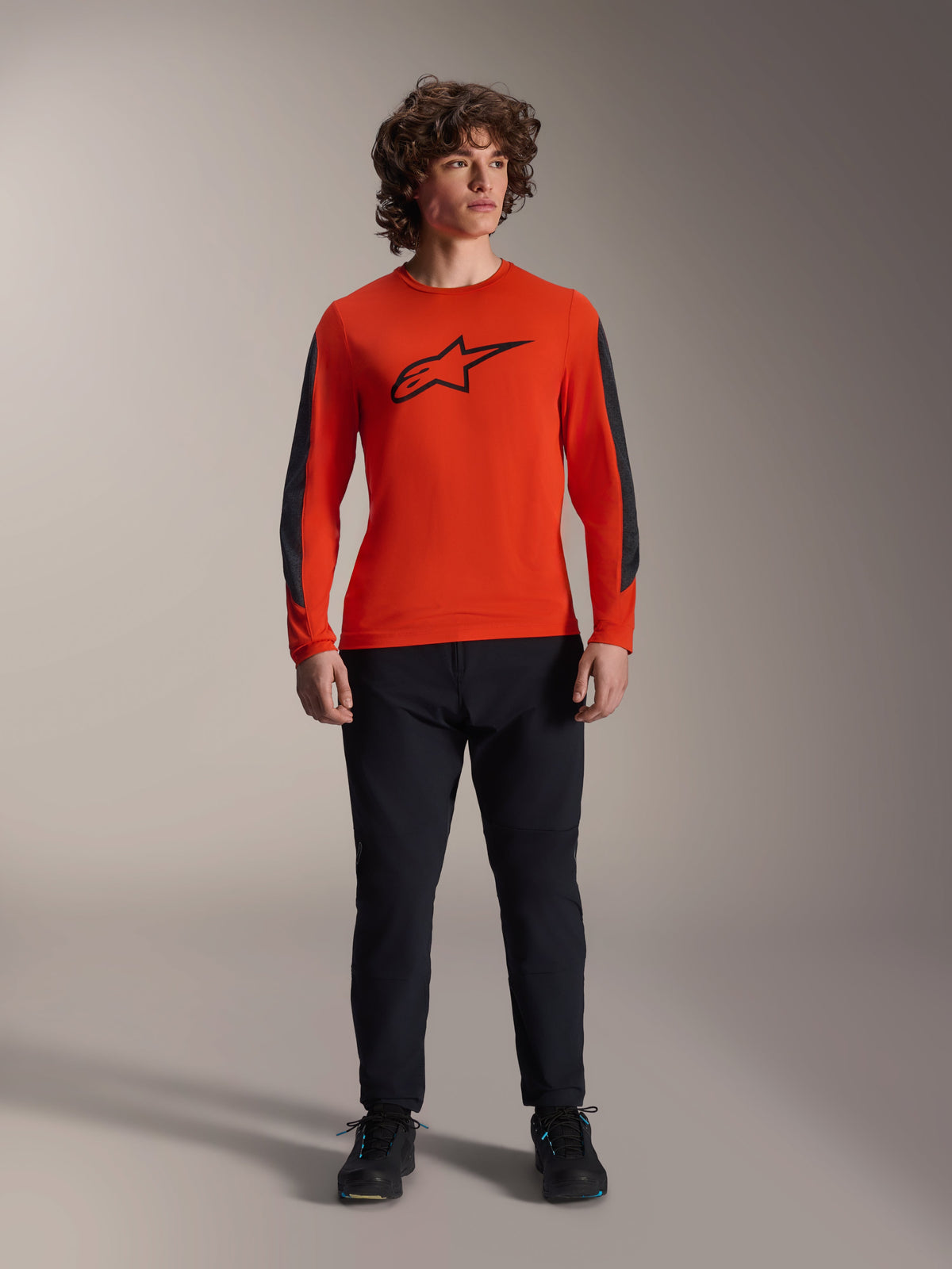 Alpinestars A-Dura Insert Astar Trikot - Langarm, Spicy Orange mit schwarzem Astar-Logo und dunkelgrauen Ärmeleinsätzen, getragen mit schwarzer Mountainbike-Hose und Schuhen, strapazierfähige Textilkonstruktion für Offroad-Fahrten