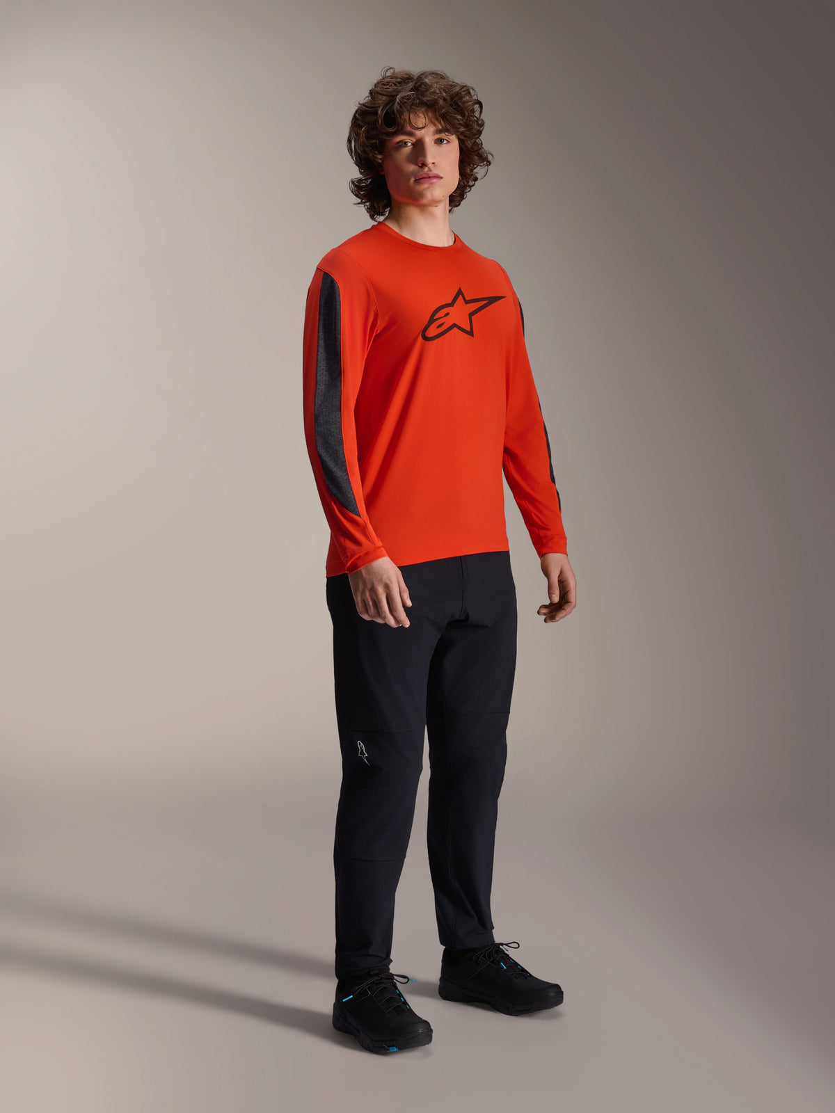 Männliches Model im Alpinestars A-Dura Insert Astar Langarm-Trikot, Spicy Orange mit schwarzem Astar-Logo auf der Brust, dunkelgrauen Ärmeleinsätzen, kombiniert mit einer schwarzen Hose, professionelle MTB-Radsportbekleidung.