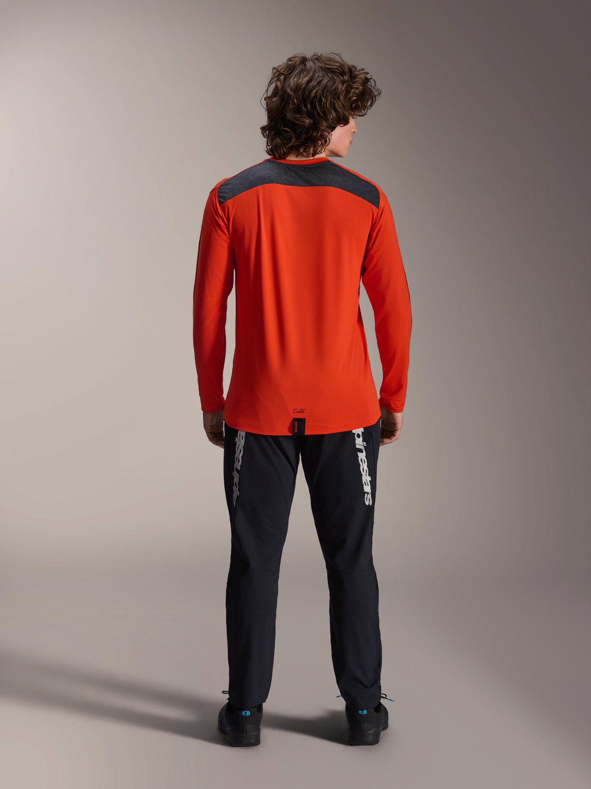Alpinestars A-Dura Insert Astar Langarm-Trikot in Spicy Orange, Rückansicht mit anthrazitgrauem Schulterpanel, getragen mit schwarzer Alpinestars Hose und schwarzen Schuhen, professionelle Studioaufnahme für MTB-Performance-Ausrüstung