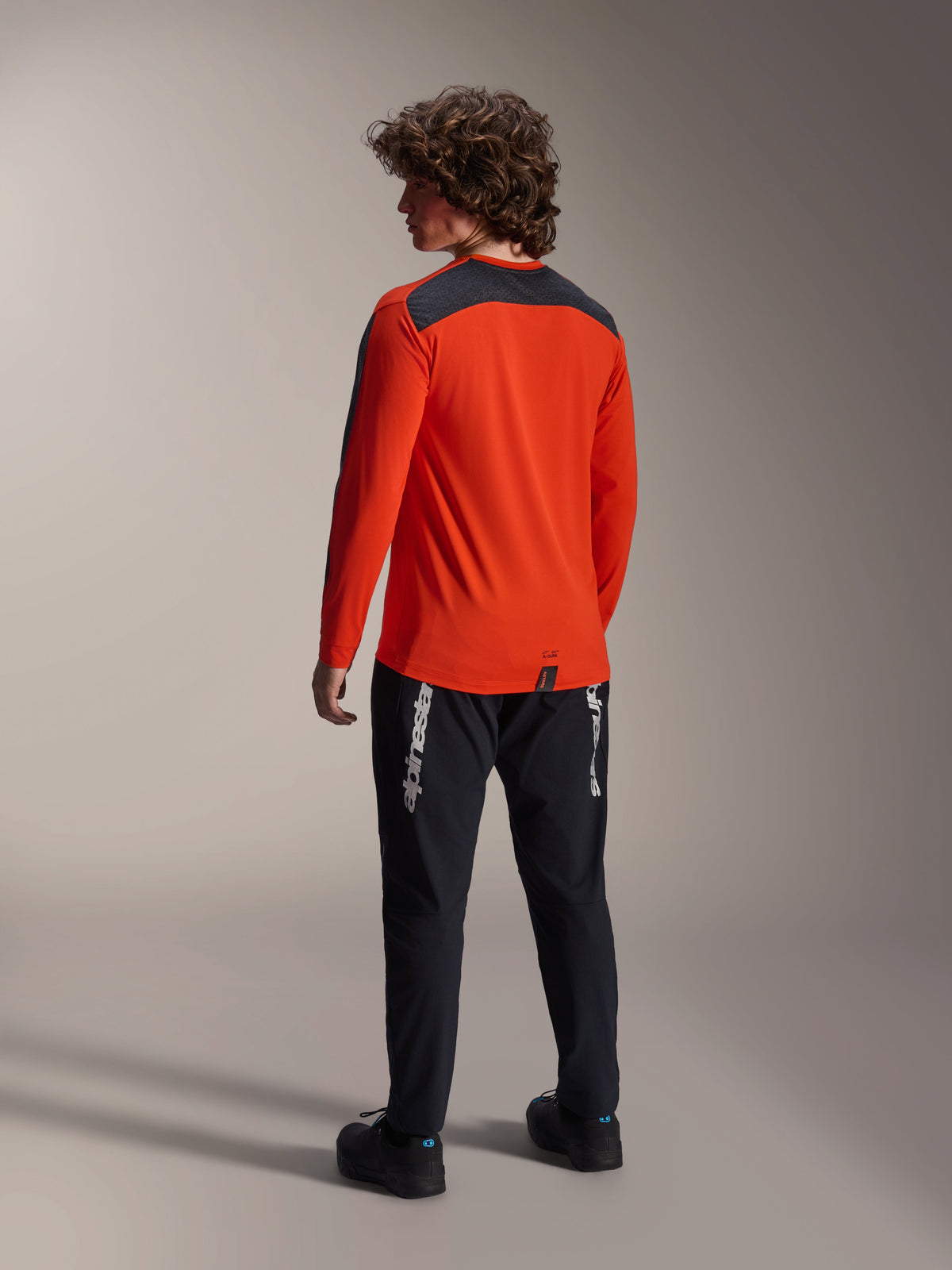 Rückansicht eines Models im Alpinestars A-Dura Insert Astar Trikot in Spicy Orange, langärmeliges MTB-Trikot mit dunkelgrauem Schultereinsatz, kombiniert mit einer schwarzen Markenhose, Offroad-Radsportausrüstung.