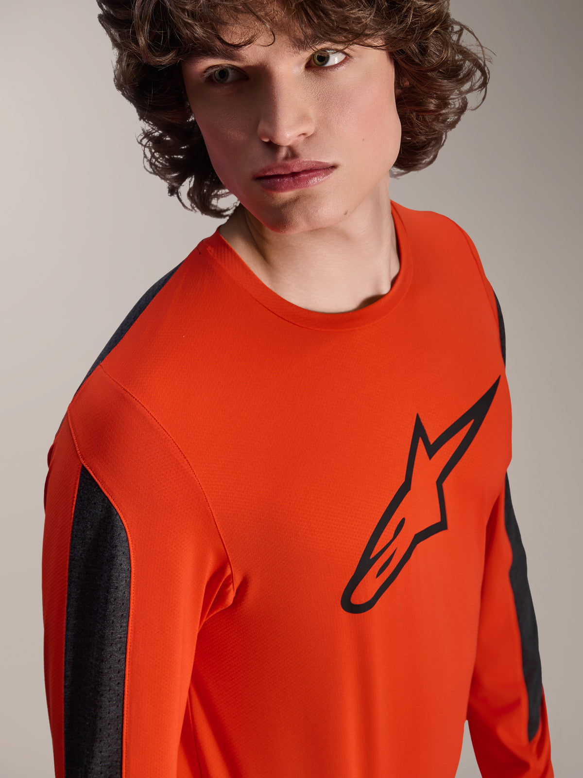 Alpinestars A-Dura Insert Astar Langarm-Trikot, MTB-Trikot in Spicy Orange mit großem schwarzem Astar-Logo auf der Brust, dunkelgraue Ärmeleinsätze, getragen von einem Model in einer Studio-Nahaufnahme, texturiertes Funktionsgewebe