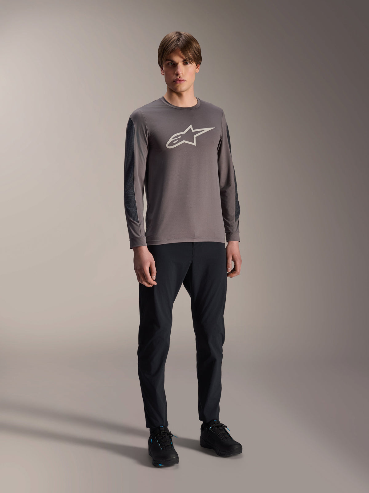 Alpinestars A-Dura Insert Astar Trikot, Langarm-MTB-Trikot, Dunkelgrau, mit großem weißem Brustlogo, getragen mit schwarzer technischer Hose und schwarzen Schuhen, Offroad-Radsportbekleidung
