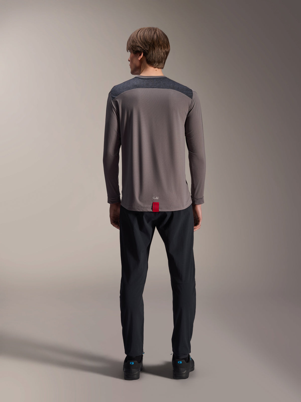 Alpinestars A-Dura Insert Astar Langarm-Trikot in Dunkelgrau, Rückansicht, mit anthrazitfarbenen Schulterpartien und einem roten Etikett am Saum, getragen mit schwarzer Hose und Schuhen, Mountainbike-Performance-Bekleidung