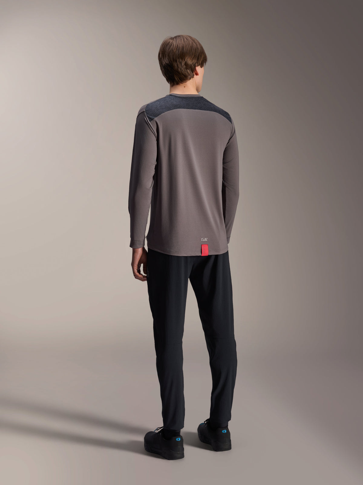 Alpinestars A-Dura Insert Astar Langarm-Trikot, Dunkelgrau, Rückansicht am Modell, mit anthrazitfarbenen verstärkten Schultereinsätzen und einem kleinen roten Etikett am Saum, kombiniert mit schwarzer Hose und Schuhen, technische MTB-Bekleidung